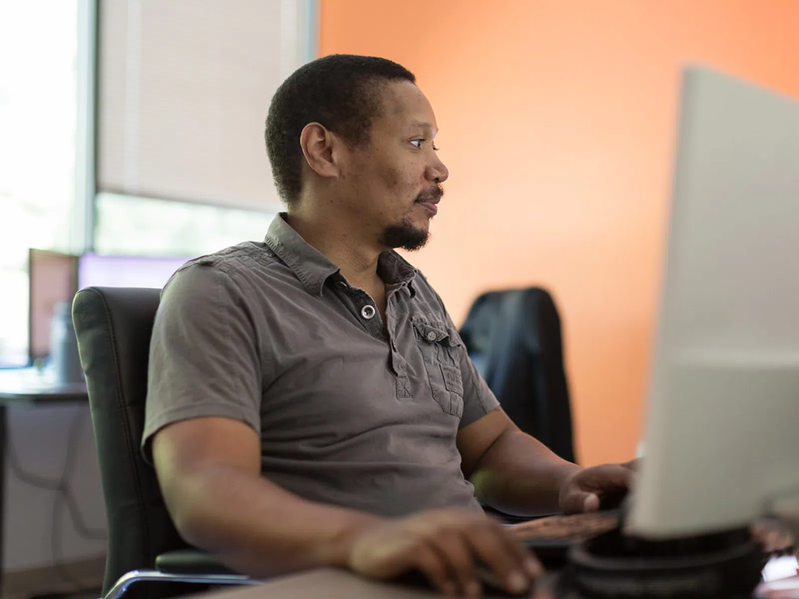 Man in gray shirt working on a desktop computer in an office with orange wall and window light.