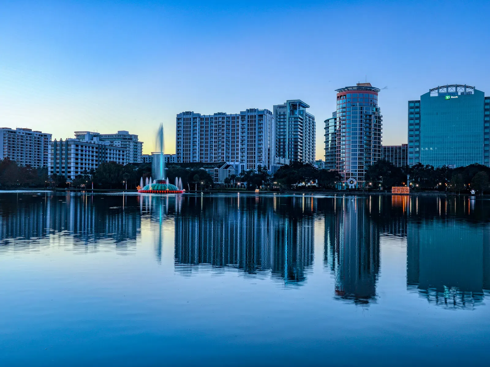 City skyline with tall buildings and central fountain reflecting on calm lake water at dusk.