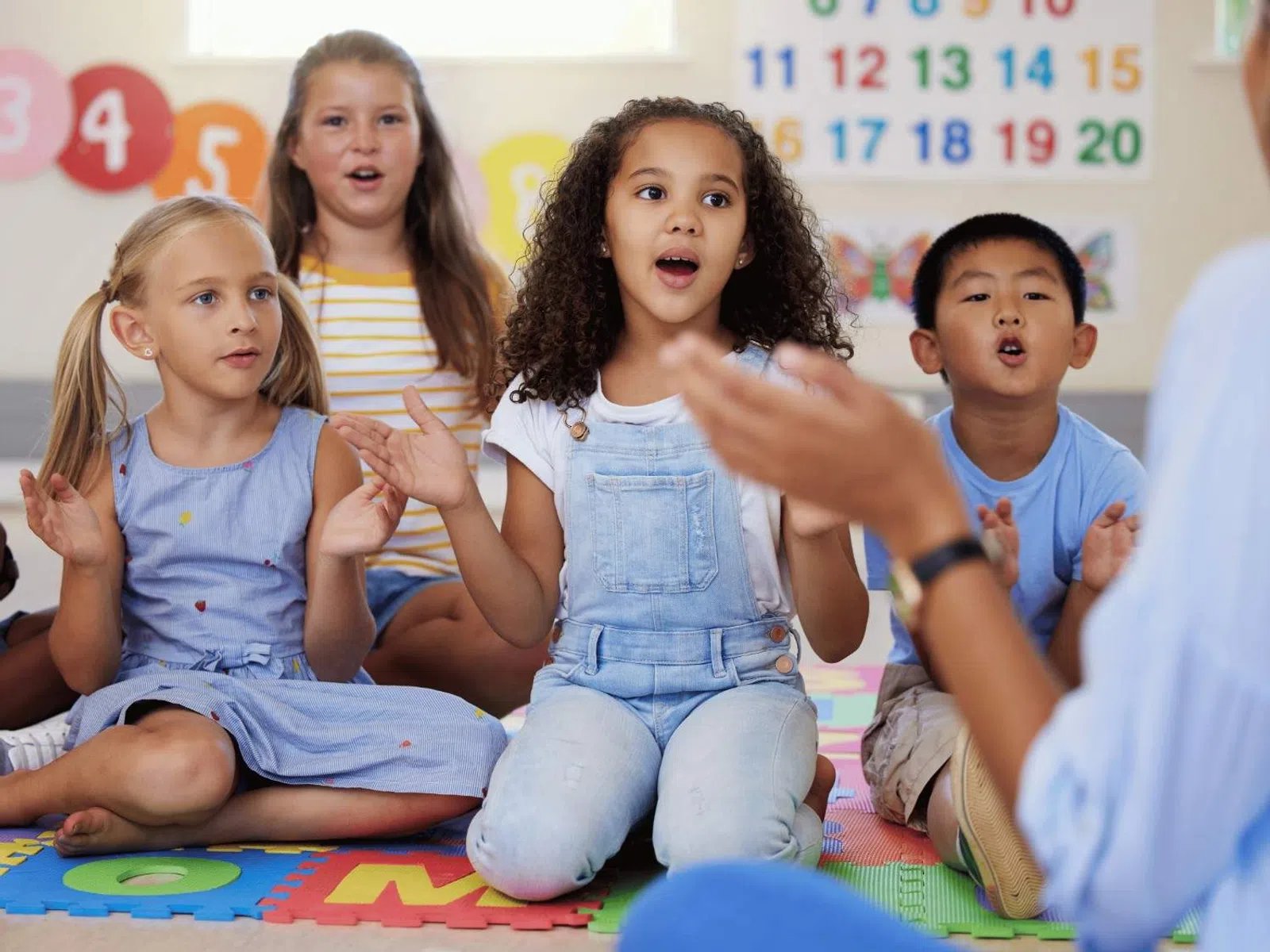 Diverse group of children sitting on a colorful mat singing and clapping in a classroom.