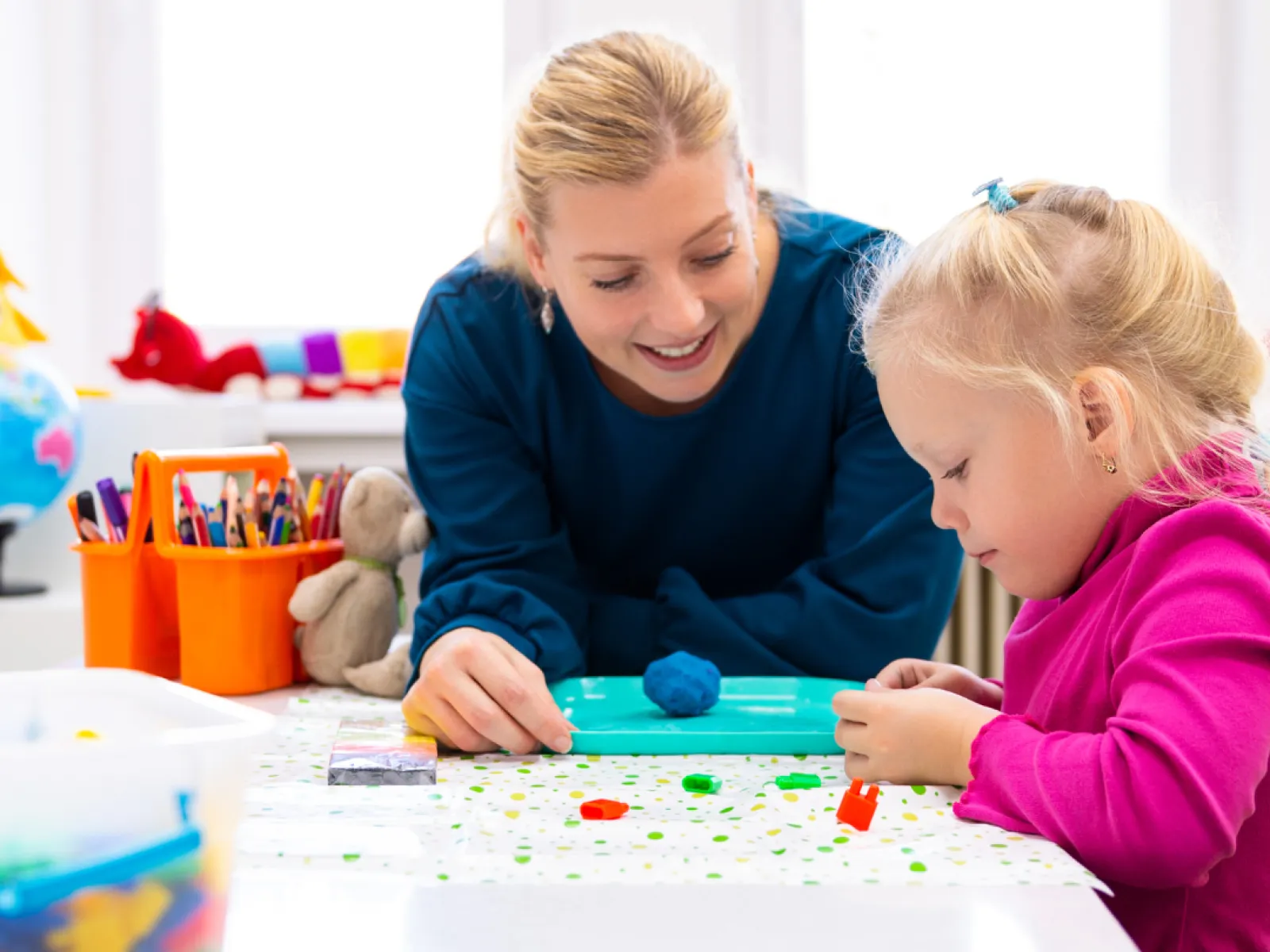 Teacher and child engaging in creative play with modeling clay and toys at a bright classroom table.