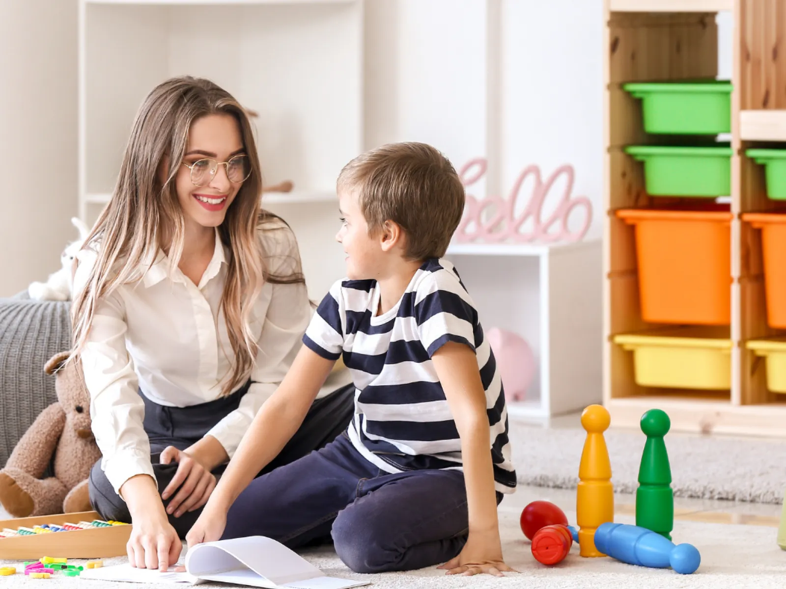 Young woman and boy sitting on floor playing and interacting in a bright, organized playroom with toys and storage bins.
