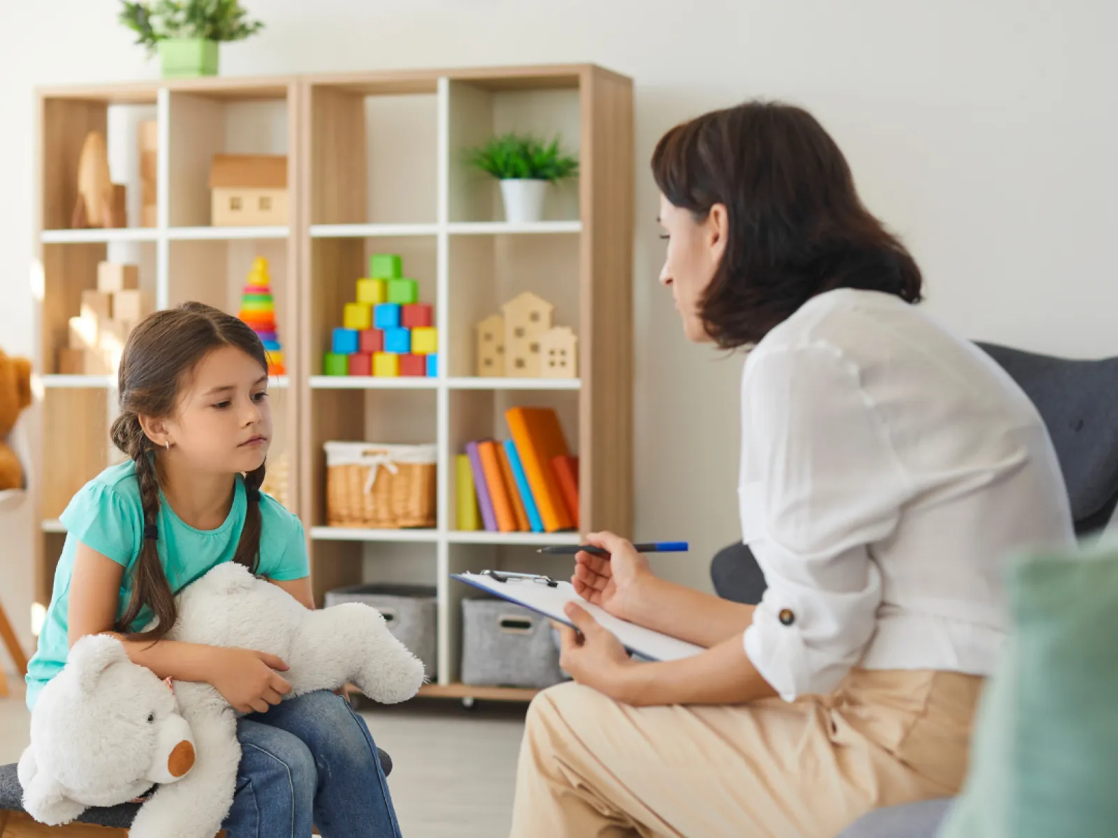 Young girl holding a teddy bear during a therapy session with a female psychologist in a cozy room.
