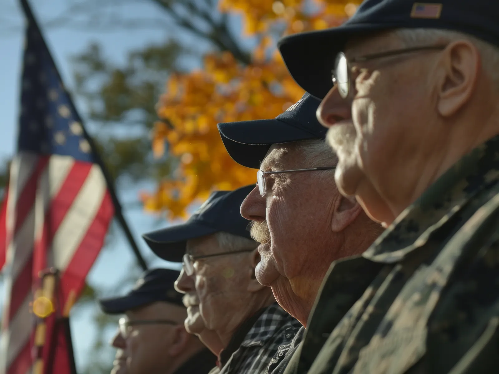 Veterans in caps stand solemnly during a ceremony with an American flag in the background.