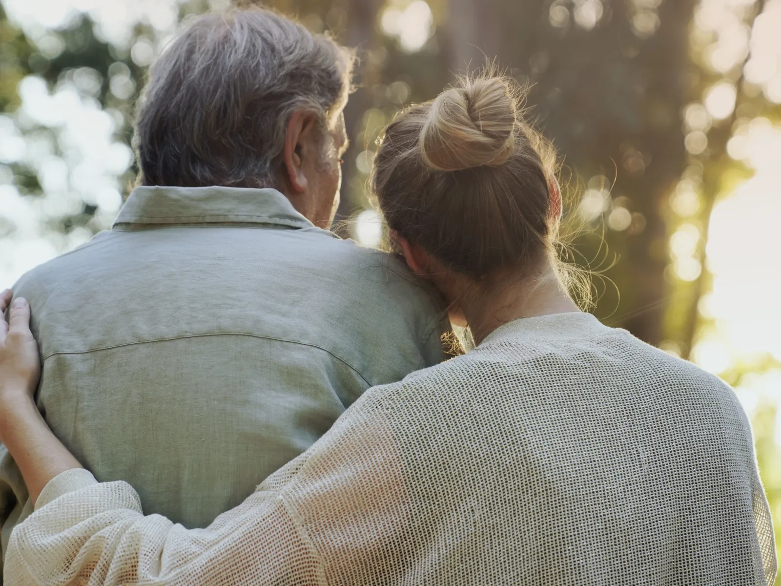 Woman hugging elderly man outdoors with sunlight filtering through trees, symbolizing care and companionship.