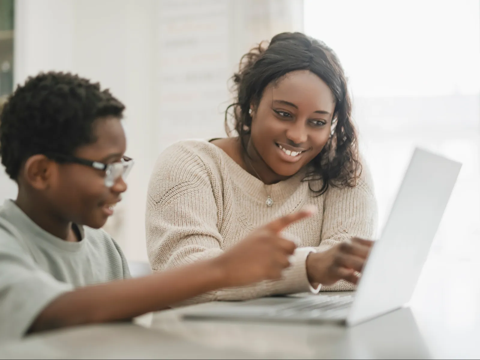 Smiling woman and young boy using laptop together in a bright room, focused on the screen and learning.