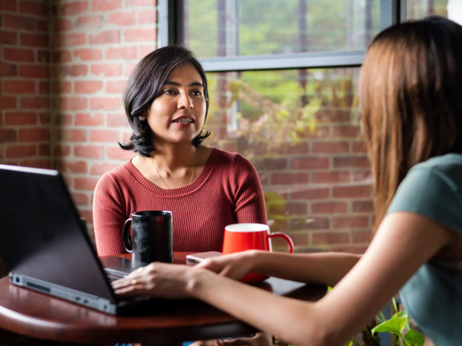 Two women having a conversation over coffee at a table with a laptop in a cozy brick-walled room with a window.