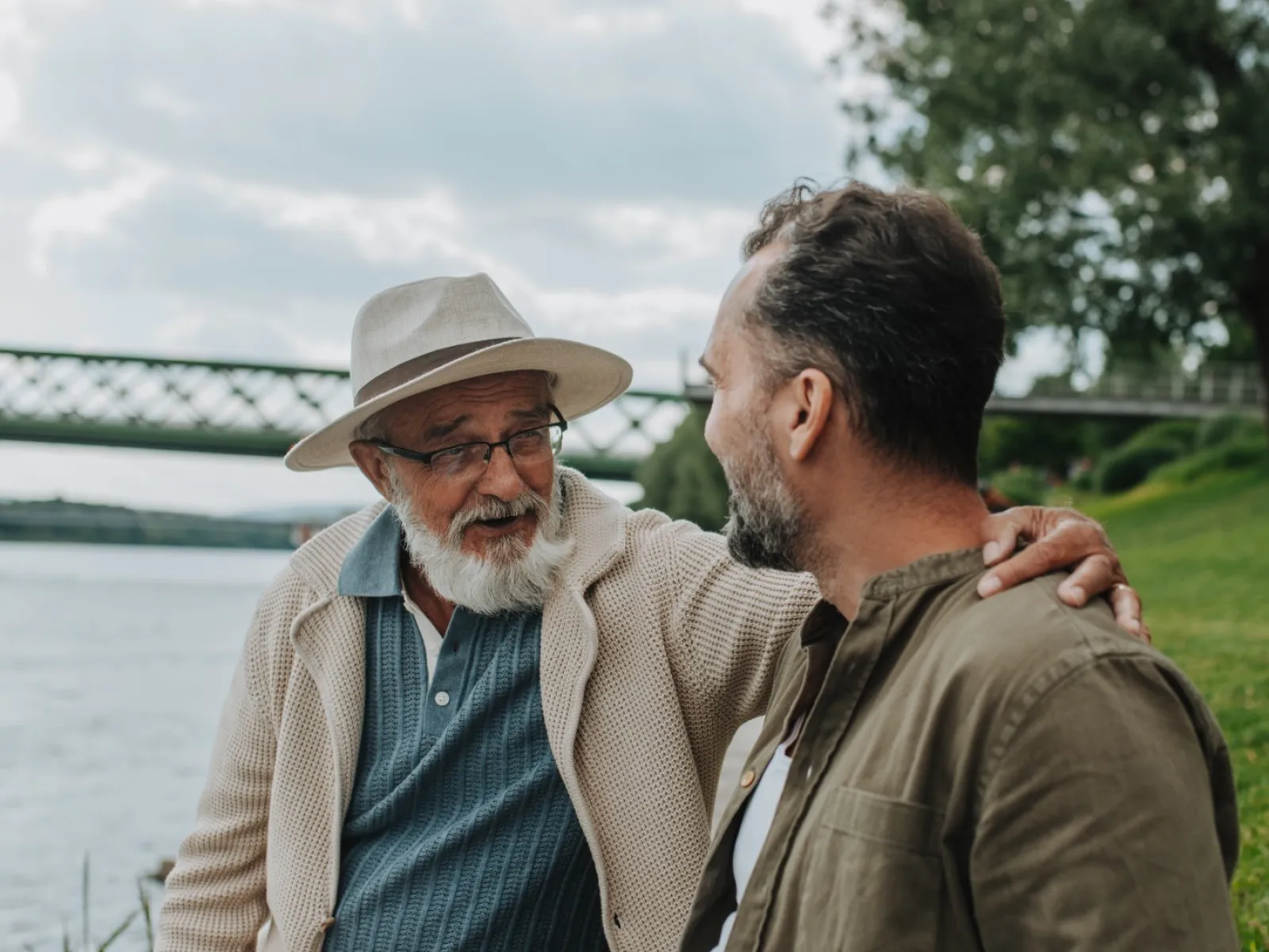 Older man with hat and glasses talking with younger man by river near green grass and bridge in background