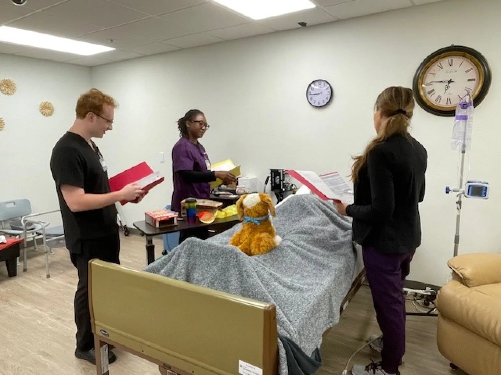 Healthcare staff review patient information in a hospital room with a covered bed and a stuffed animal.