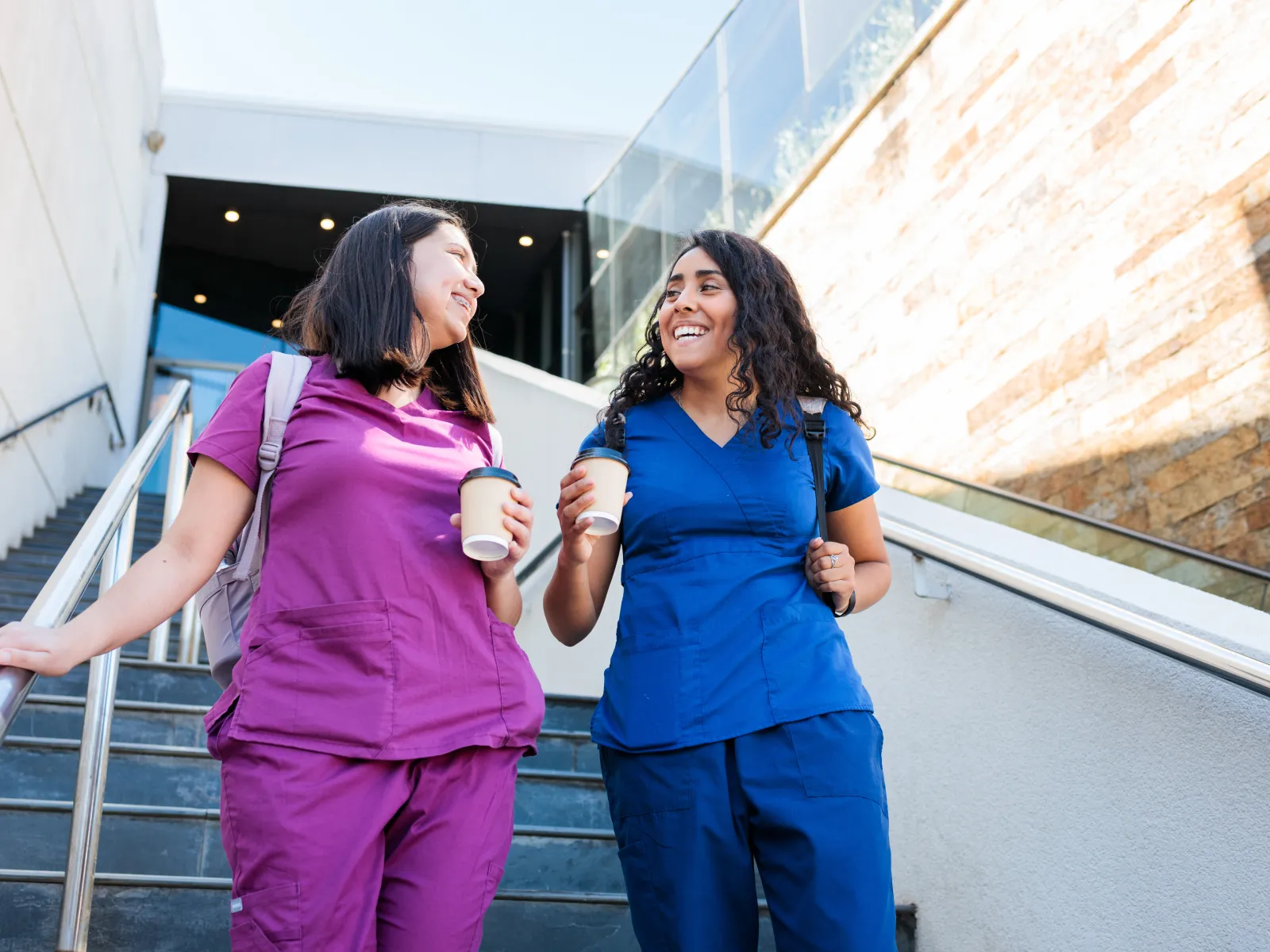 Two smiling female healthcare workers in purple and blue scrubs holding coffee cups while walking down stairs outdoors.