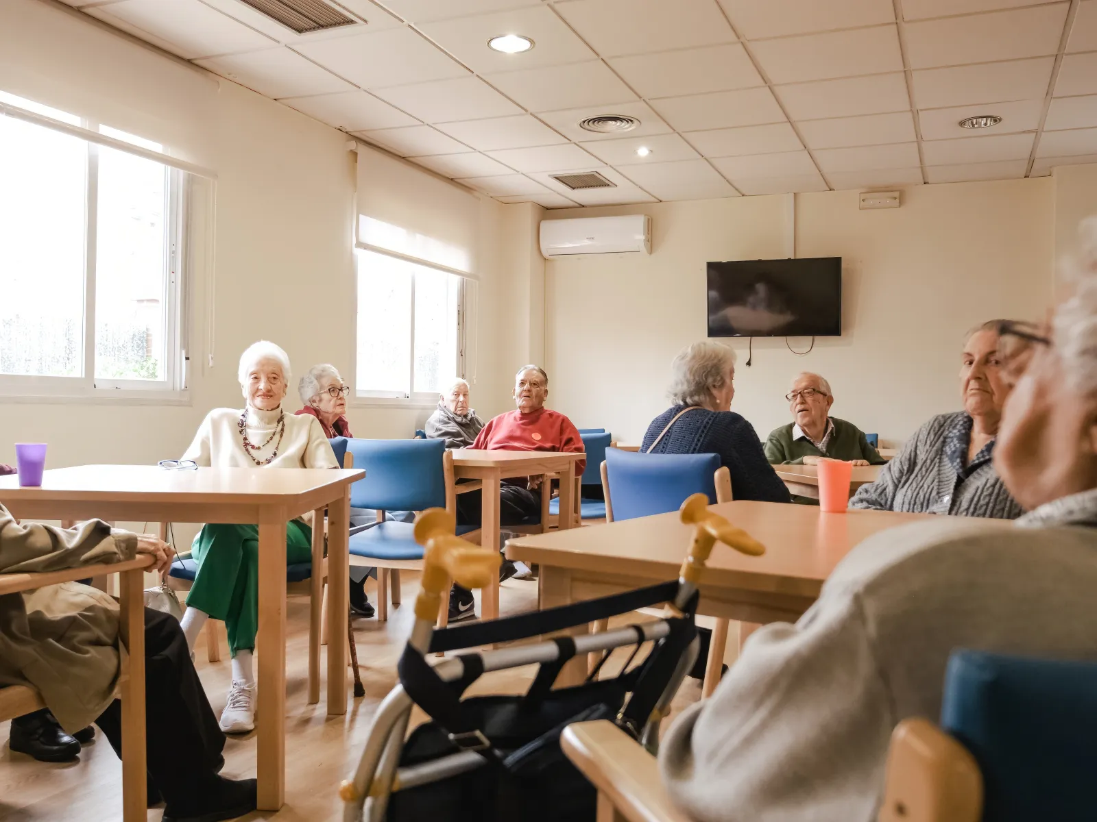 Elderly people sitting and socializing in a bright communal room with tables and chairs in a care facility.