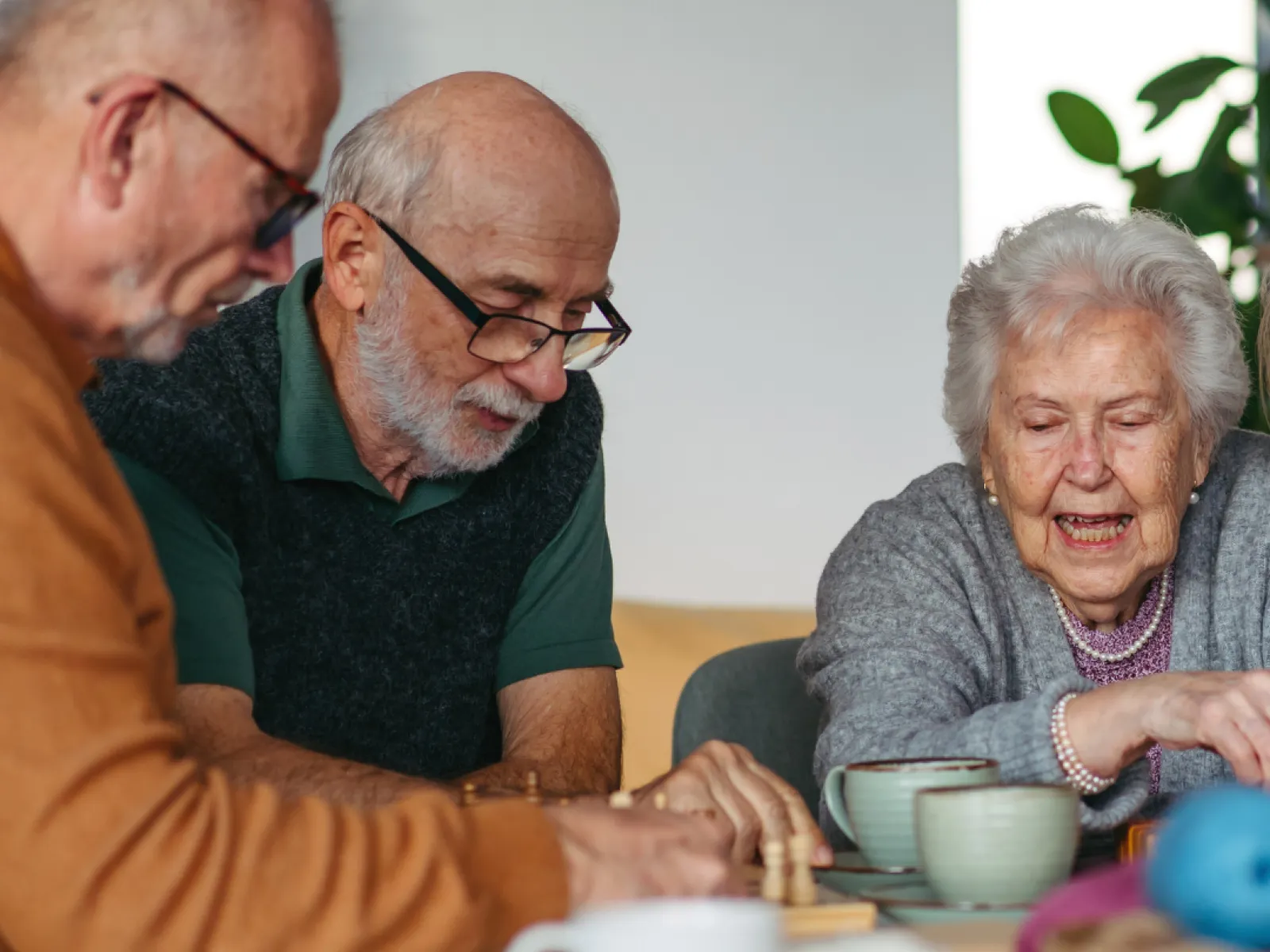 Three elderly people engaged in a board game, smiling and enjoying tea together in a cozy room.