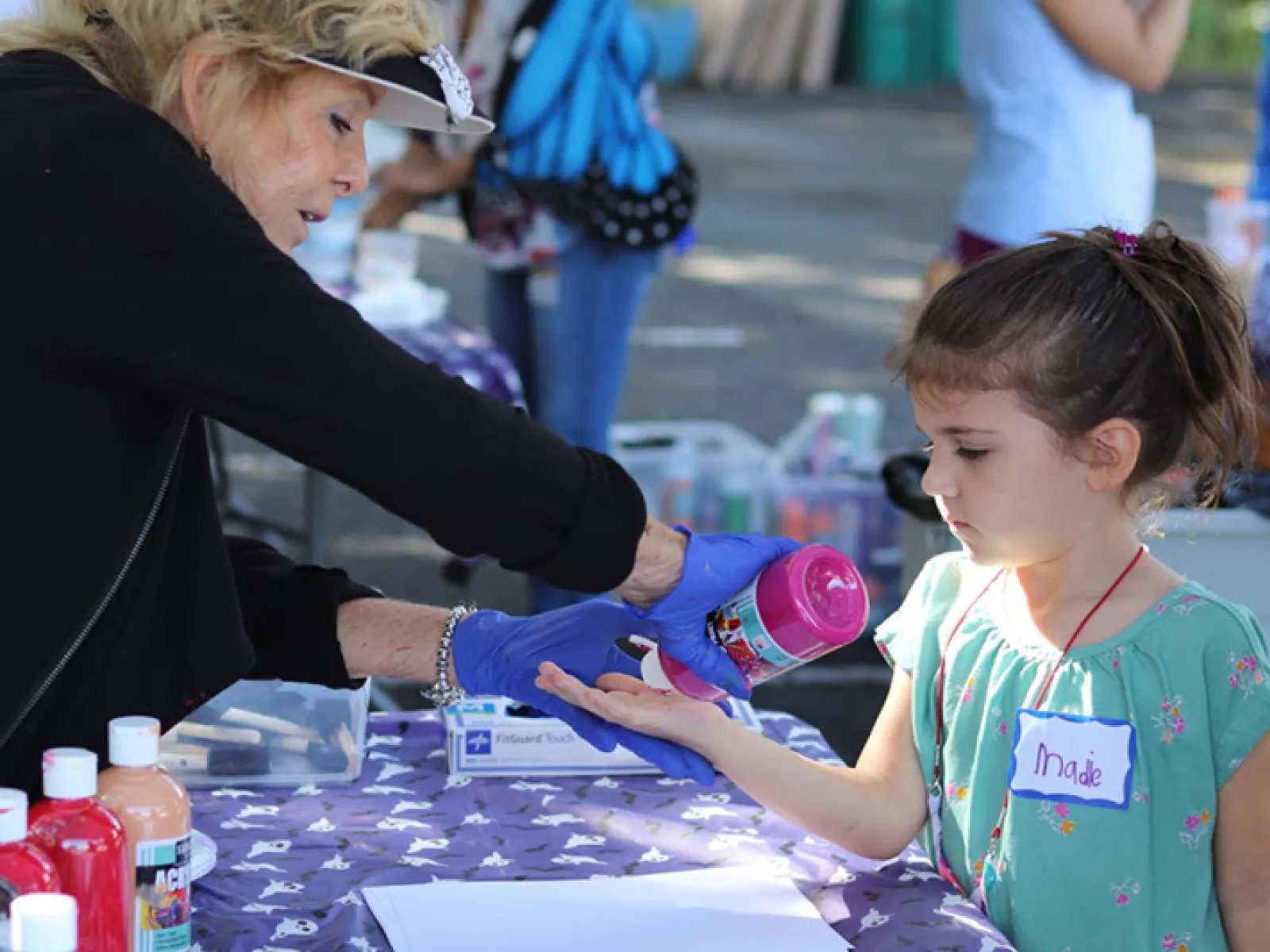 A woman applies pink paint to a young girl's hand for a craft activity at an outdoor event.