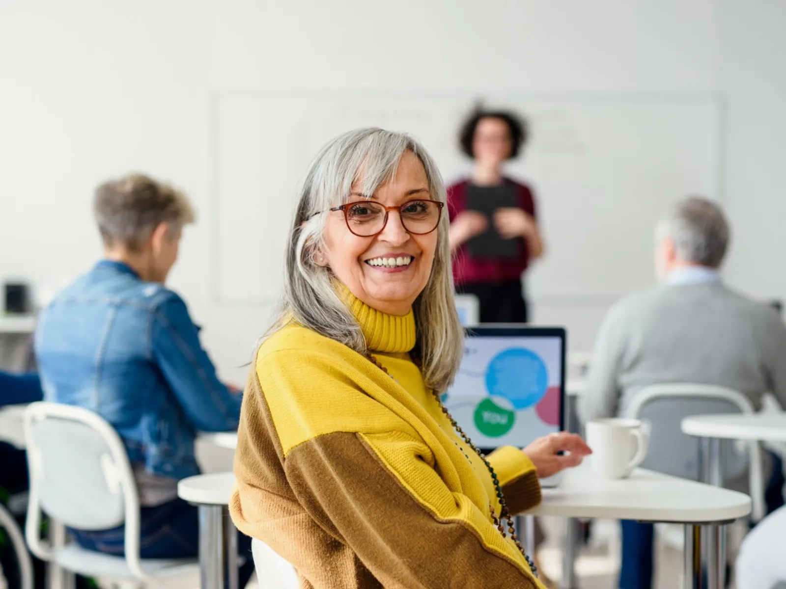 Smiling mature woman in glasses sitting at classroom table with laptop, diverse adults learning in background