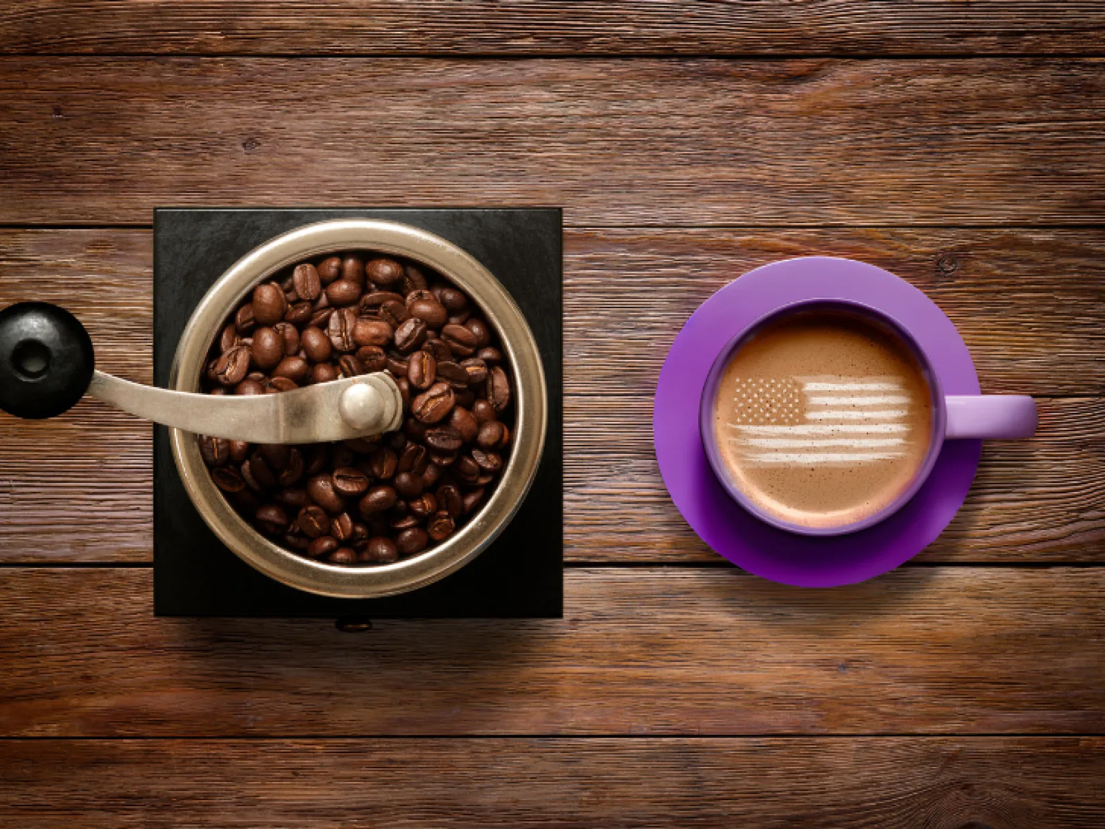 Top view of coffee grinder with beans and purple cup of coffee with American flag latte art on wooden table.