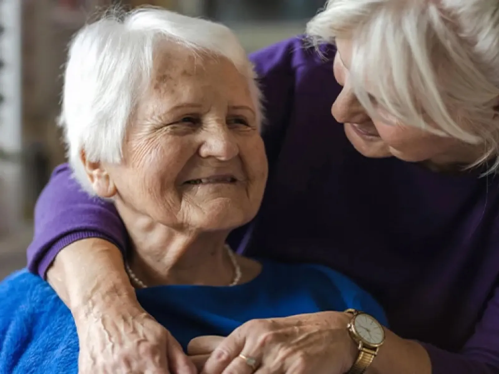 Senior woman smiling while being affectionately embraced by a loved one with white hair indoors.