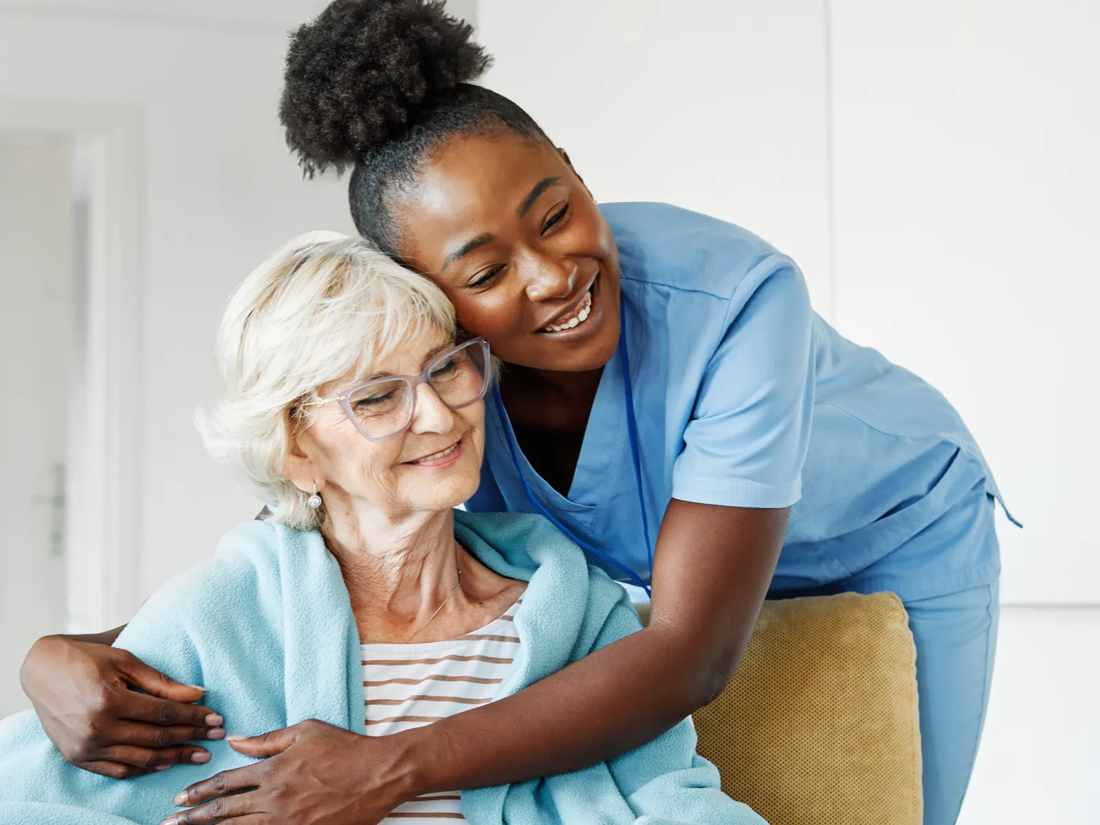 Smiling female healthcare worker hugs elderly woman in blue blanket seated in armchair at home.