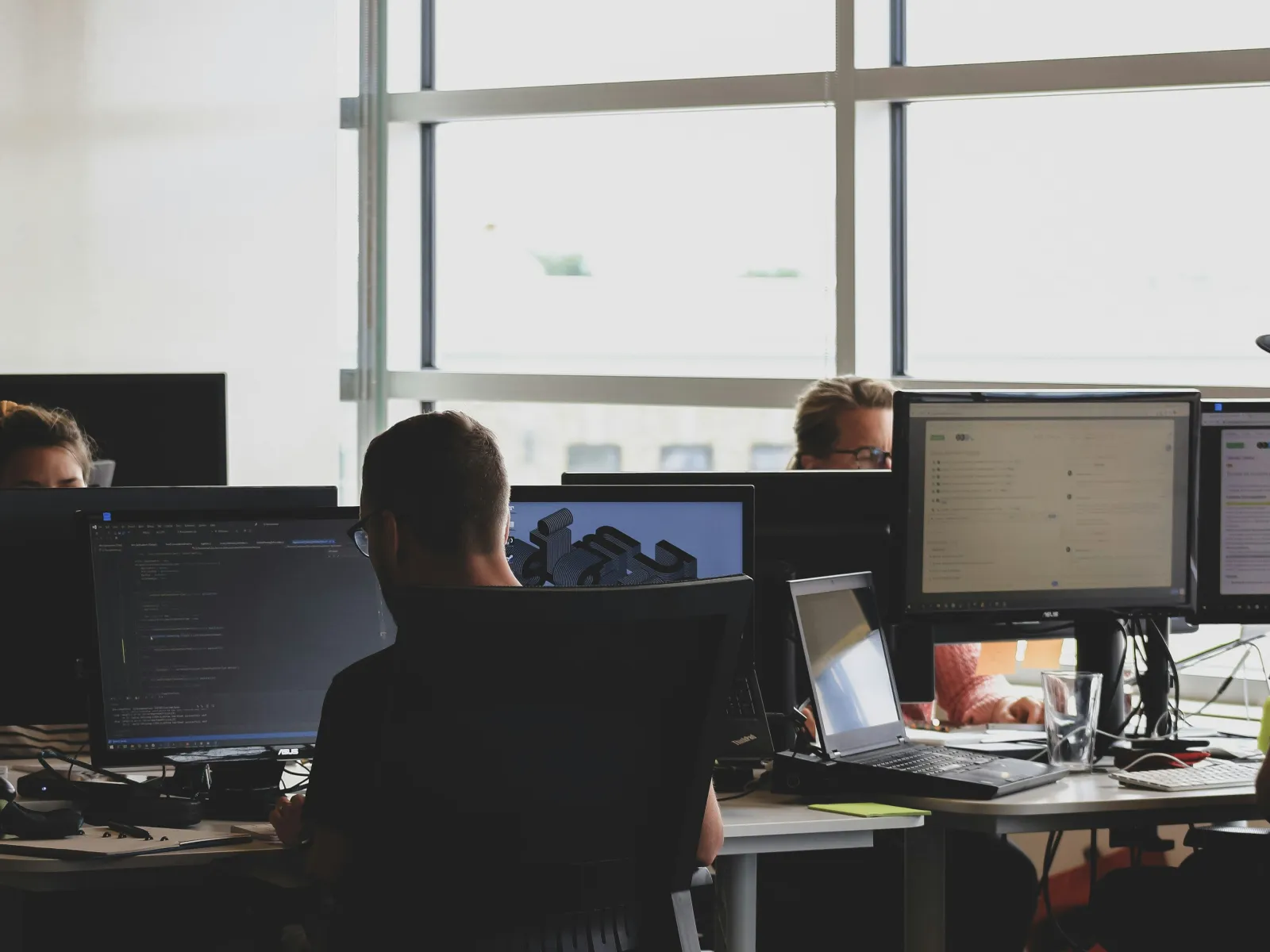 People working on computers and coding in a modern office with large windows and multiple monitors.