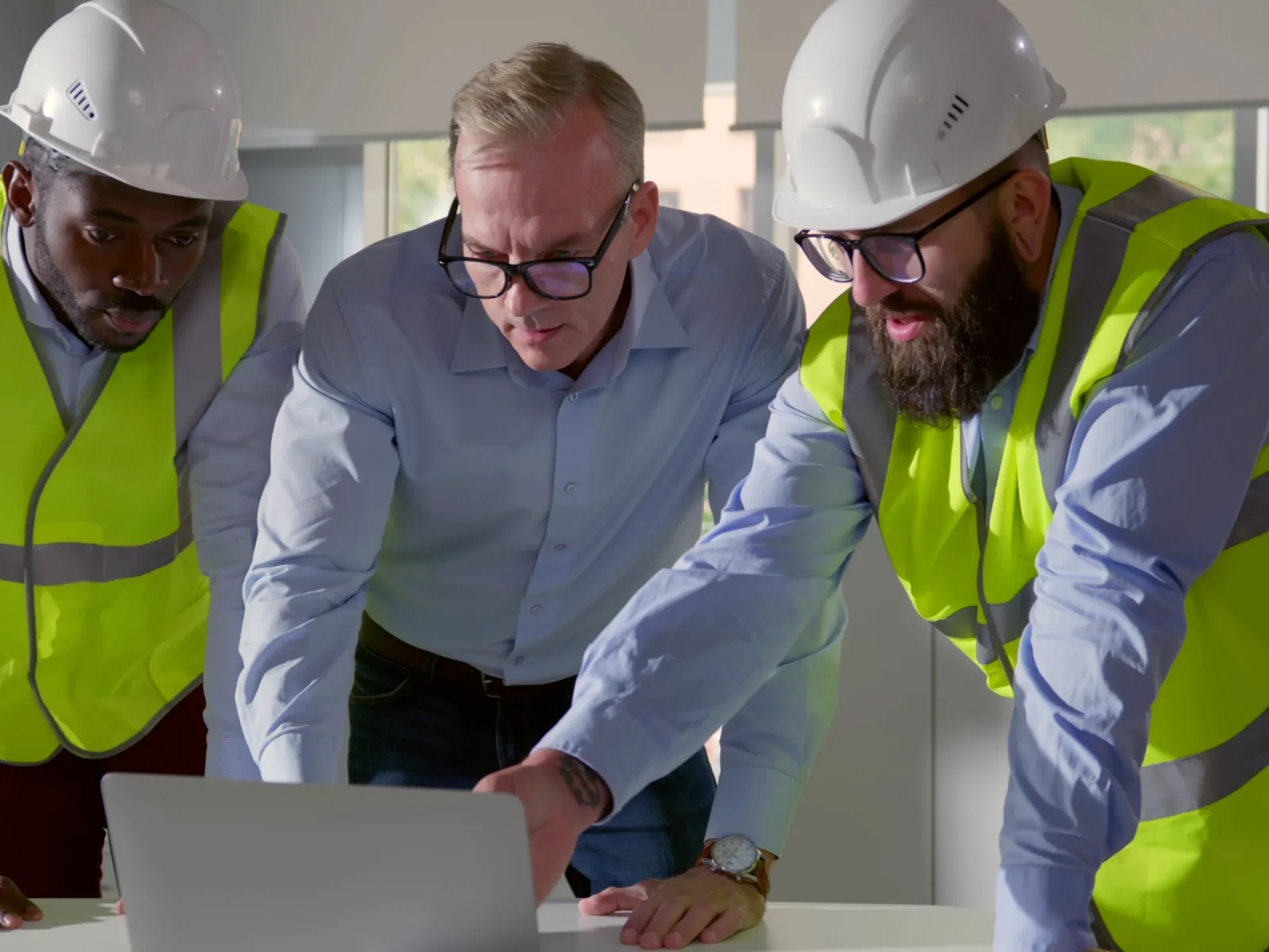 Three construction professionals in safety gear reviewing plans on a laptop in a bright office environment.
