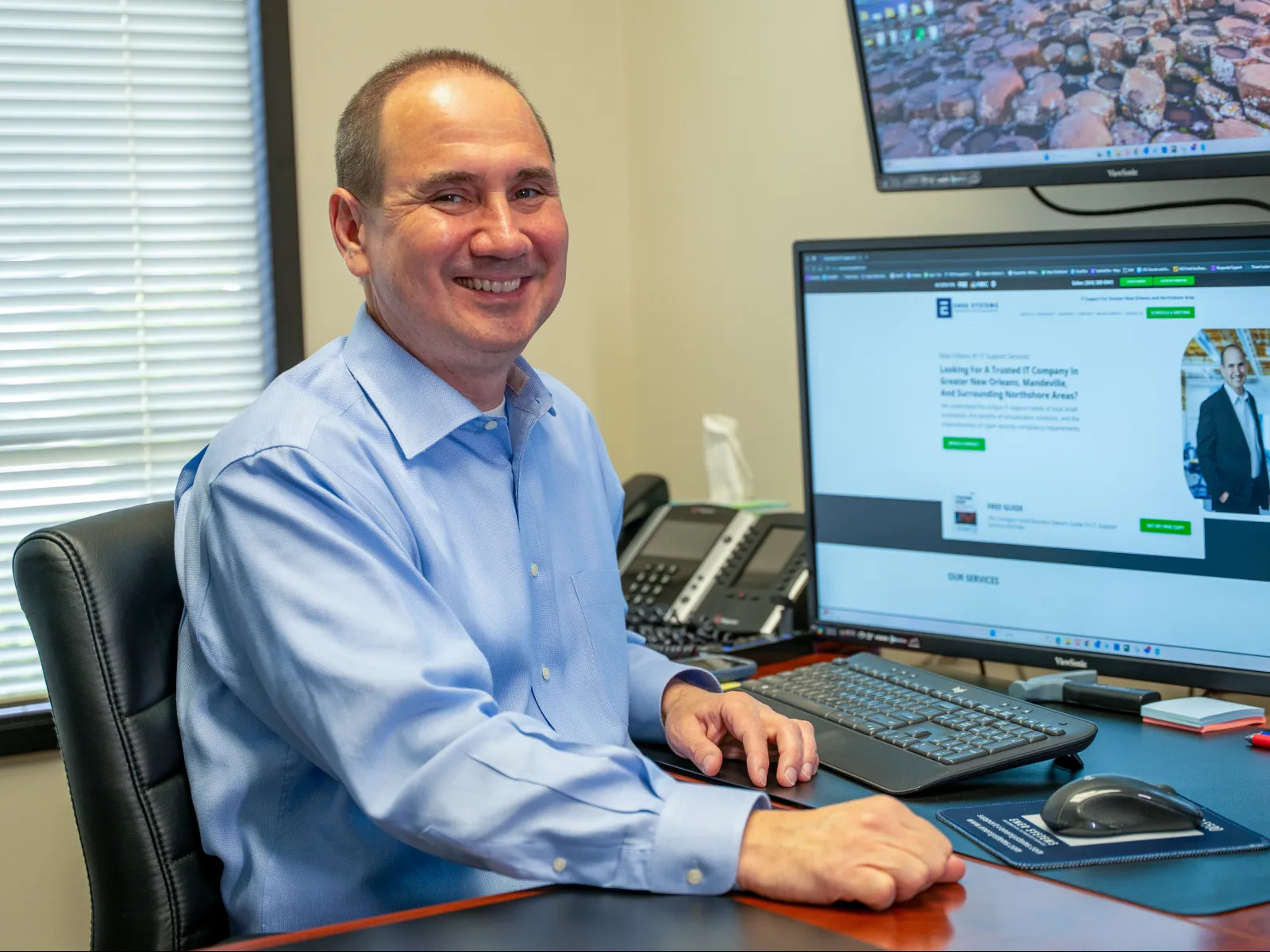 Smiling man in blue shirt sitting at desk in office with three computer monitors and office phone.