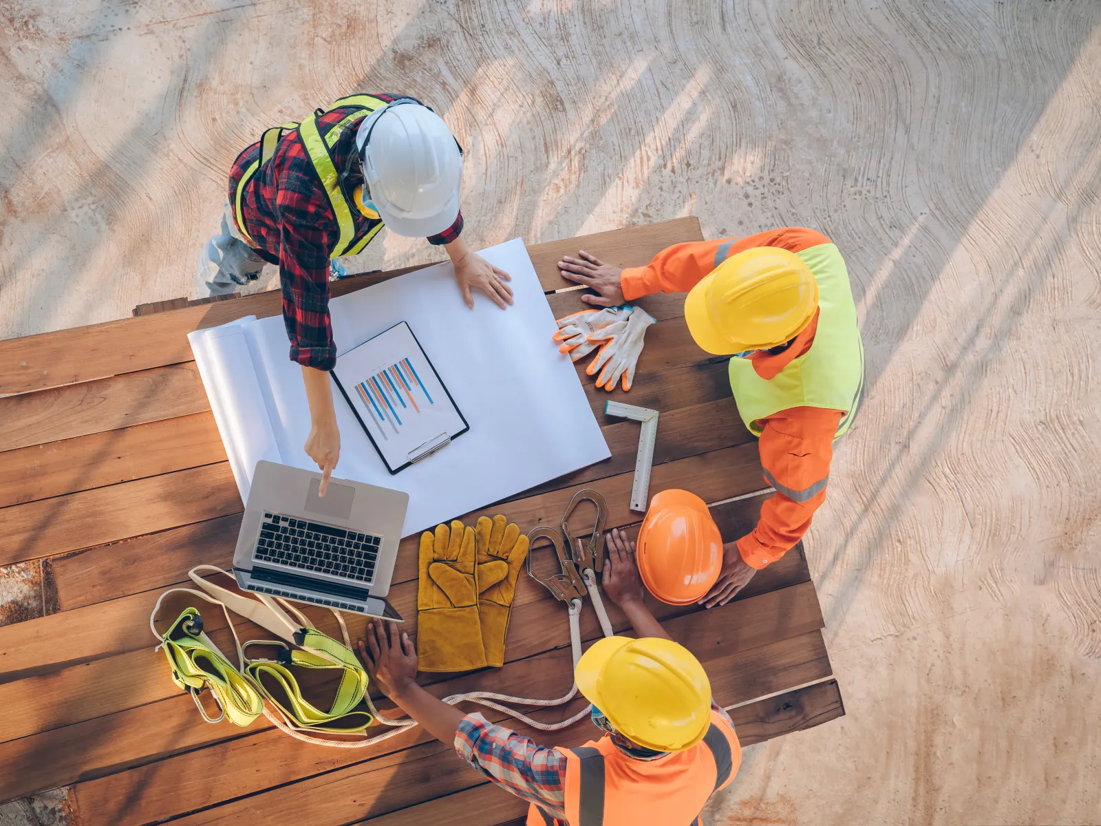 Construction workers reviewing blueprints and laptop on a wooden table with tools and safety gear outdoors.