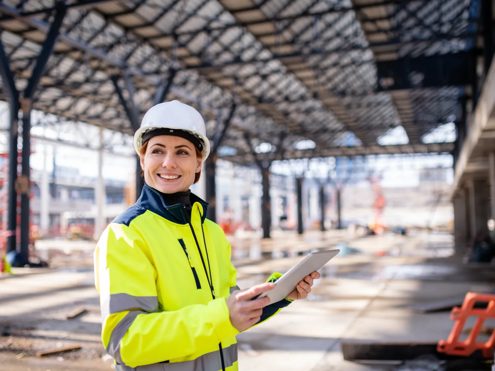 Smiling female construction worker in high-visibility jacket and hard hat holding a tablet at a building site.
