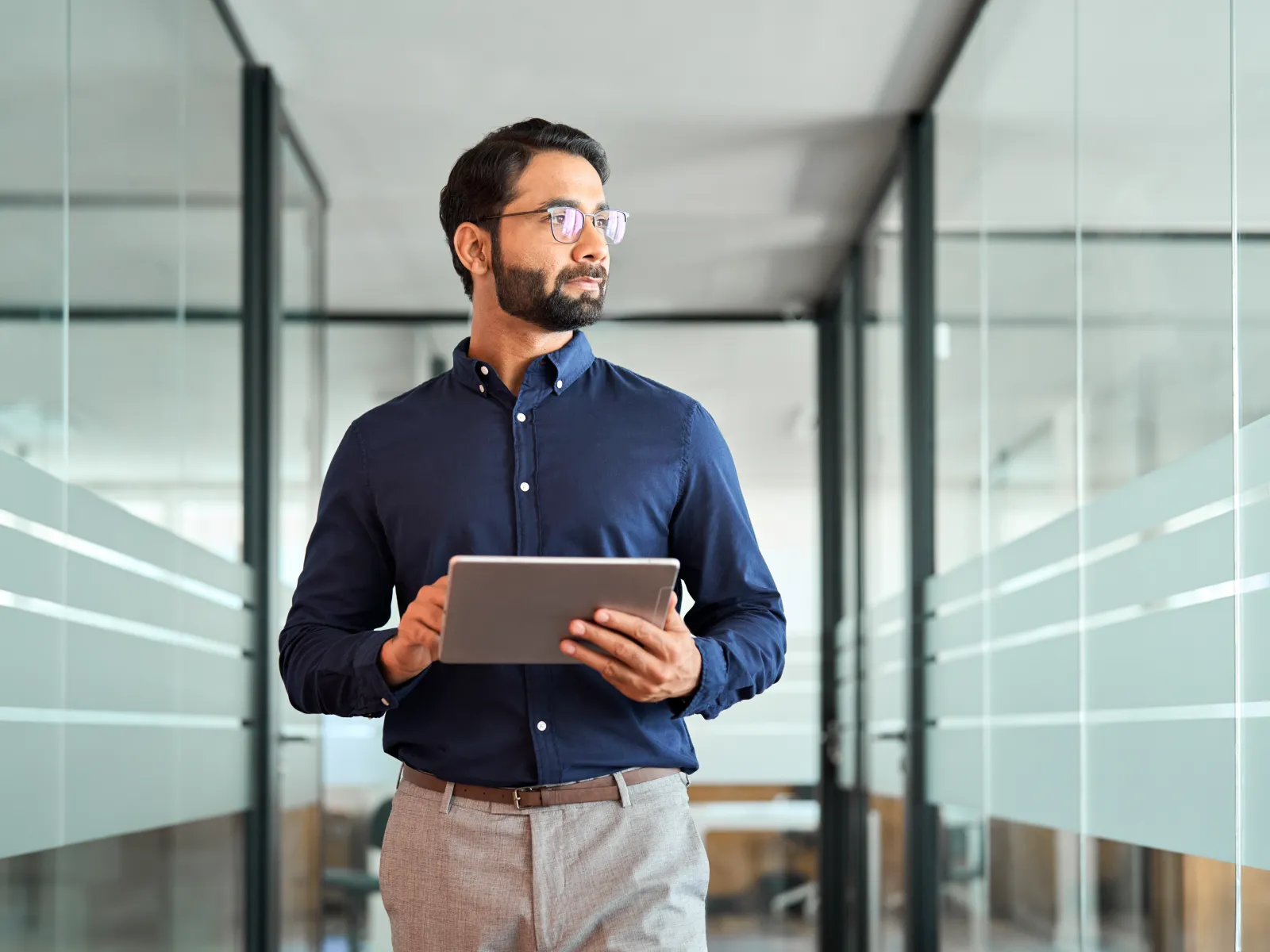 Confident businessman holding tablet walking through modern glass office corridor looking away thoughtfully
