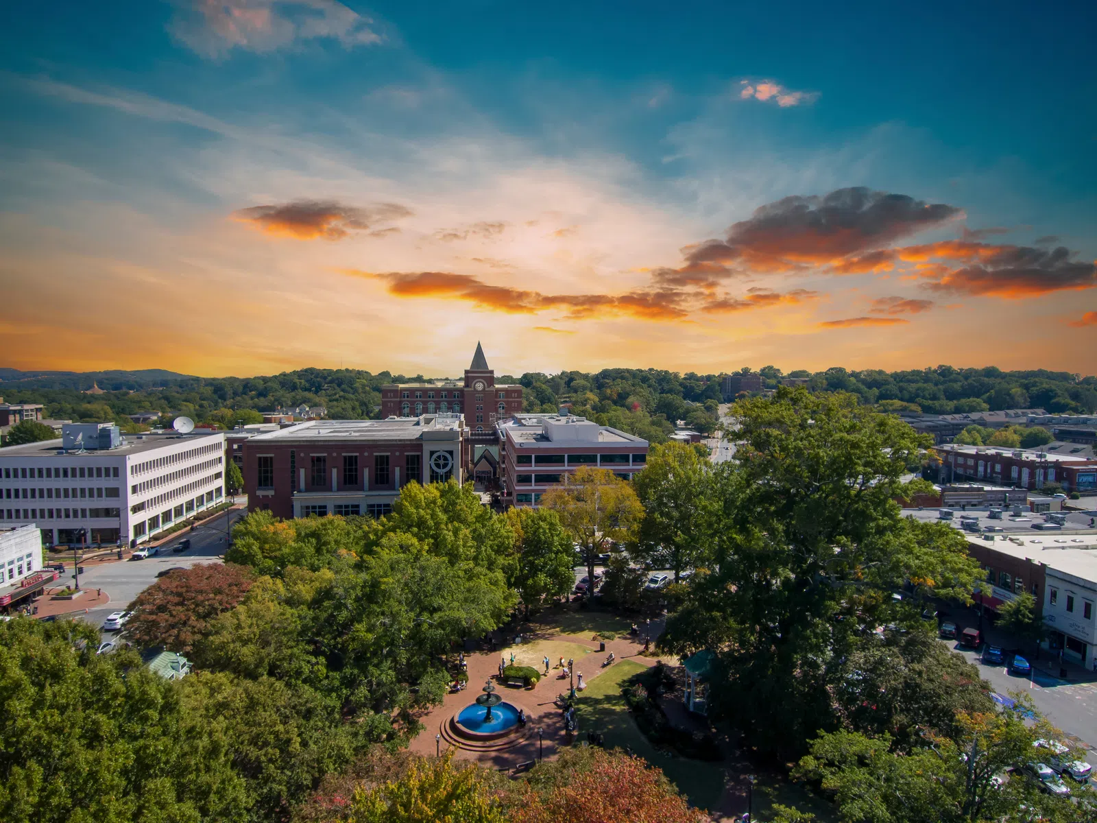 Aerial view of a town square with a fountain, surrounding trees, and buildings under a sunset sky.