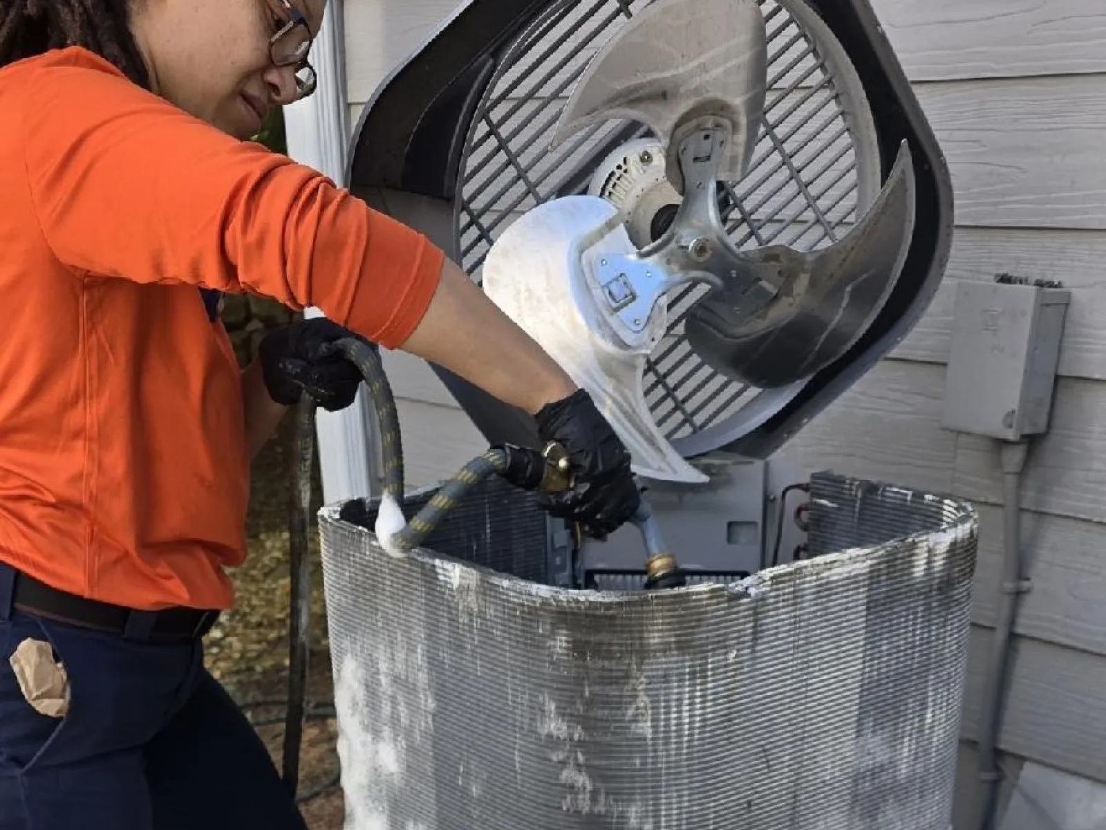 Estes Women Technician cleaning outdoor air conditioning unit with foam and hose near house exterior.