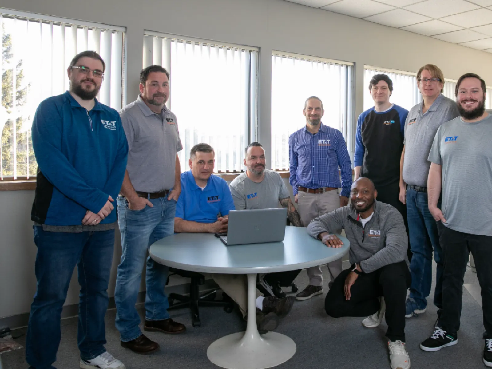 Group of professionals in casual attire gathered around a table with a laptop in an office setting.