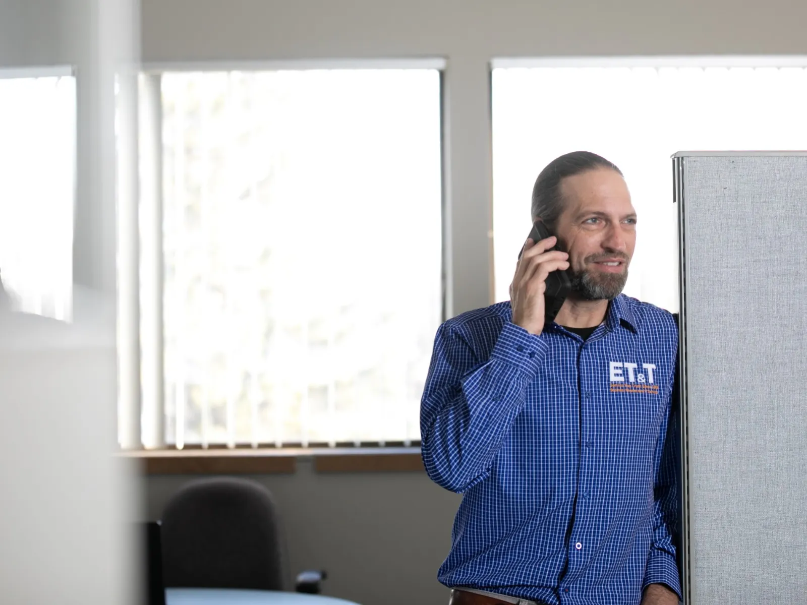 Man in blue checkered shirt talking on phone in bright office cubicle with windows and reflections.