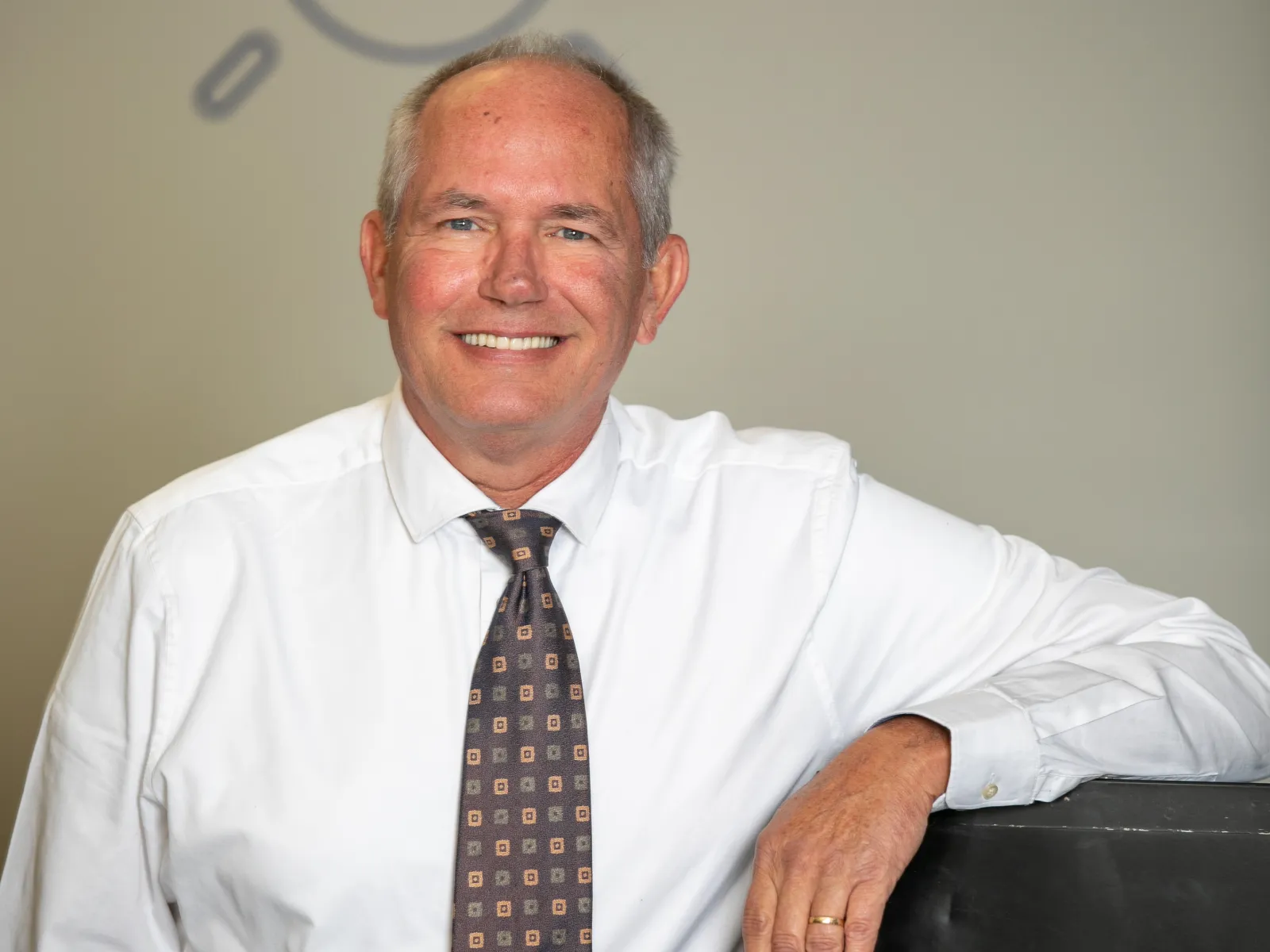 Smiling man in white shirt and patterned tie leaning on black object with lightbulb illustrations on wall.