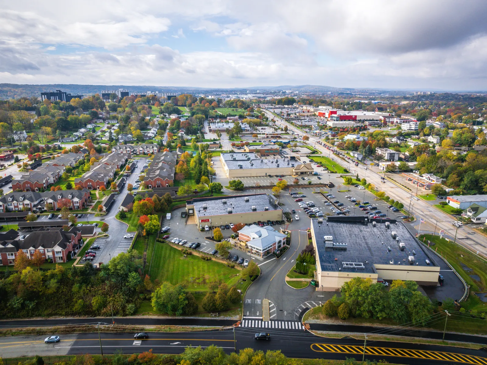 Aerial view of a suburban area featuring diverse buildings, parking lots, and lush greenery under a cloudy sky.