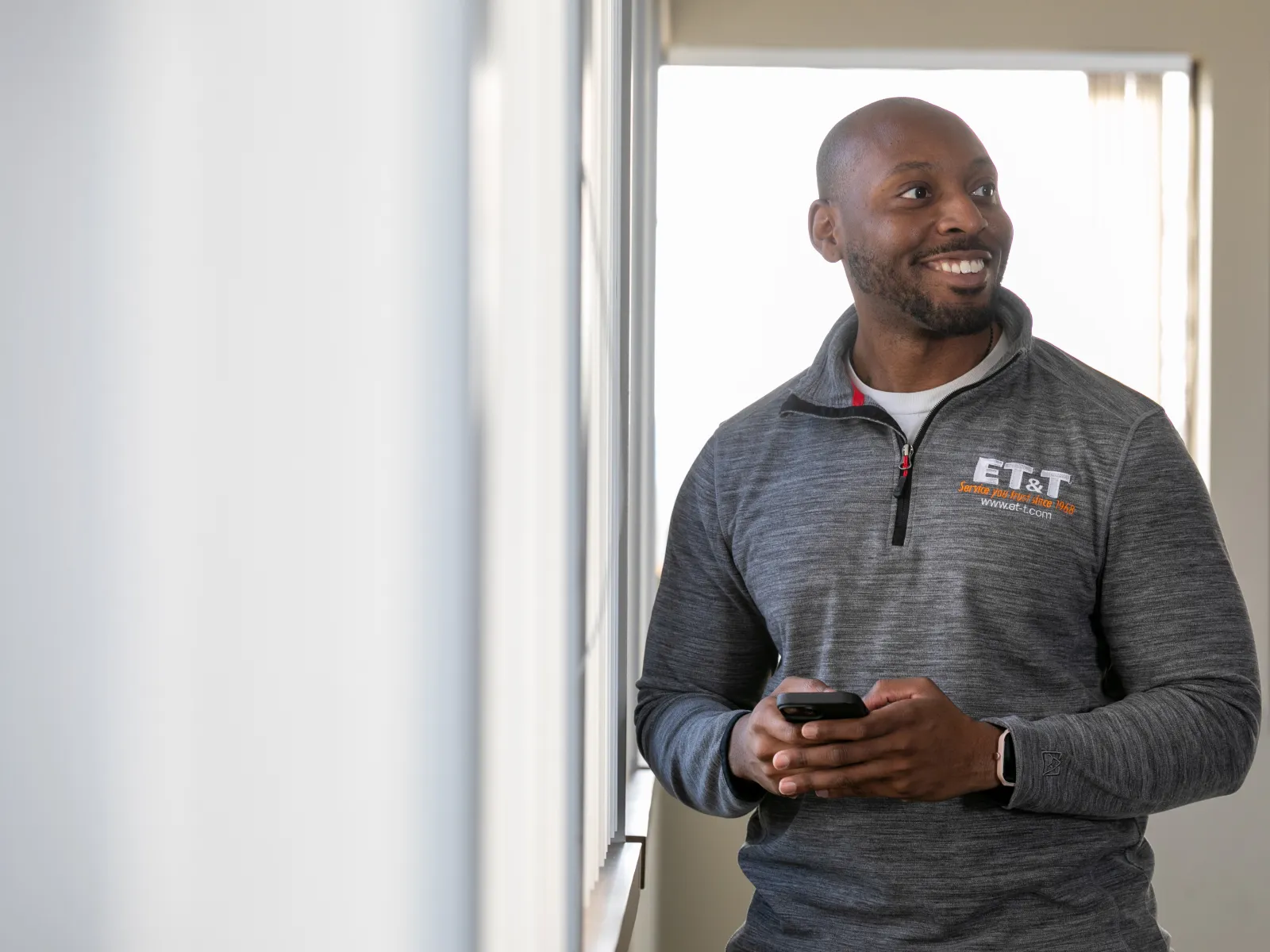 Smiling man in gray ET&T sweatshirt holding phone, looking out window in bright office space.