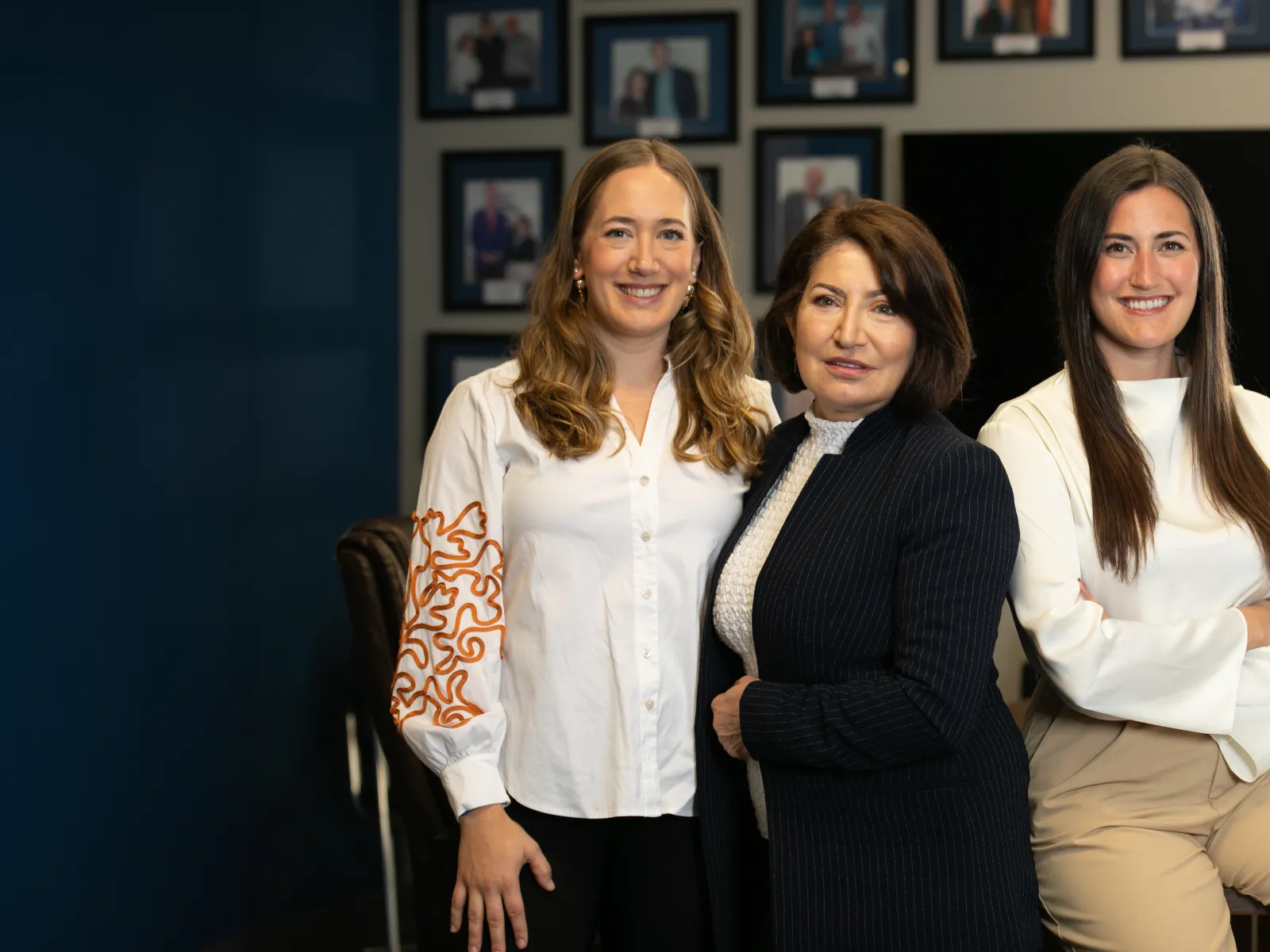 Three women professionals standing confidently in an office setting with framed photos in the background.