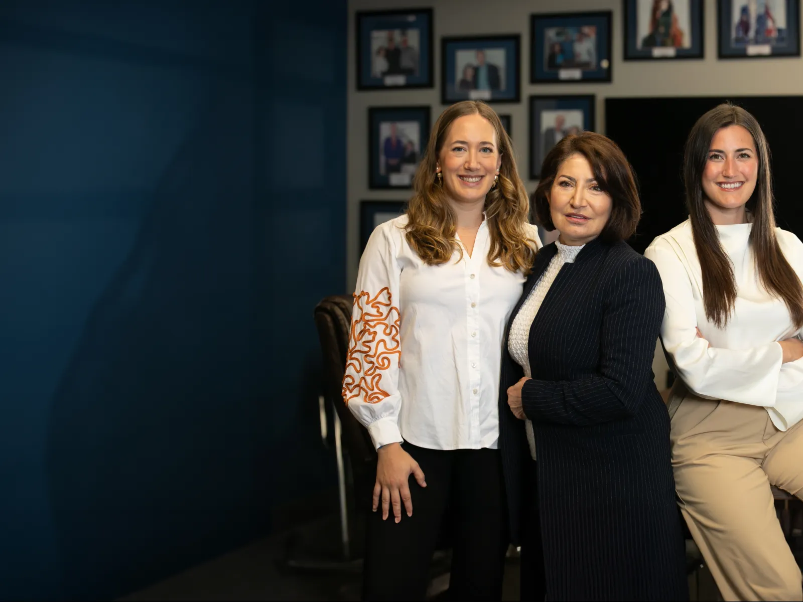 Three professional women smiling and posing in an office with framed photos on the wall behind them