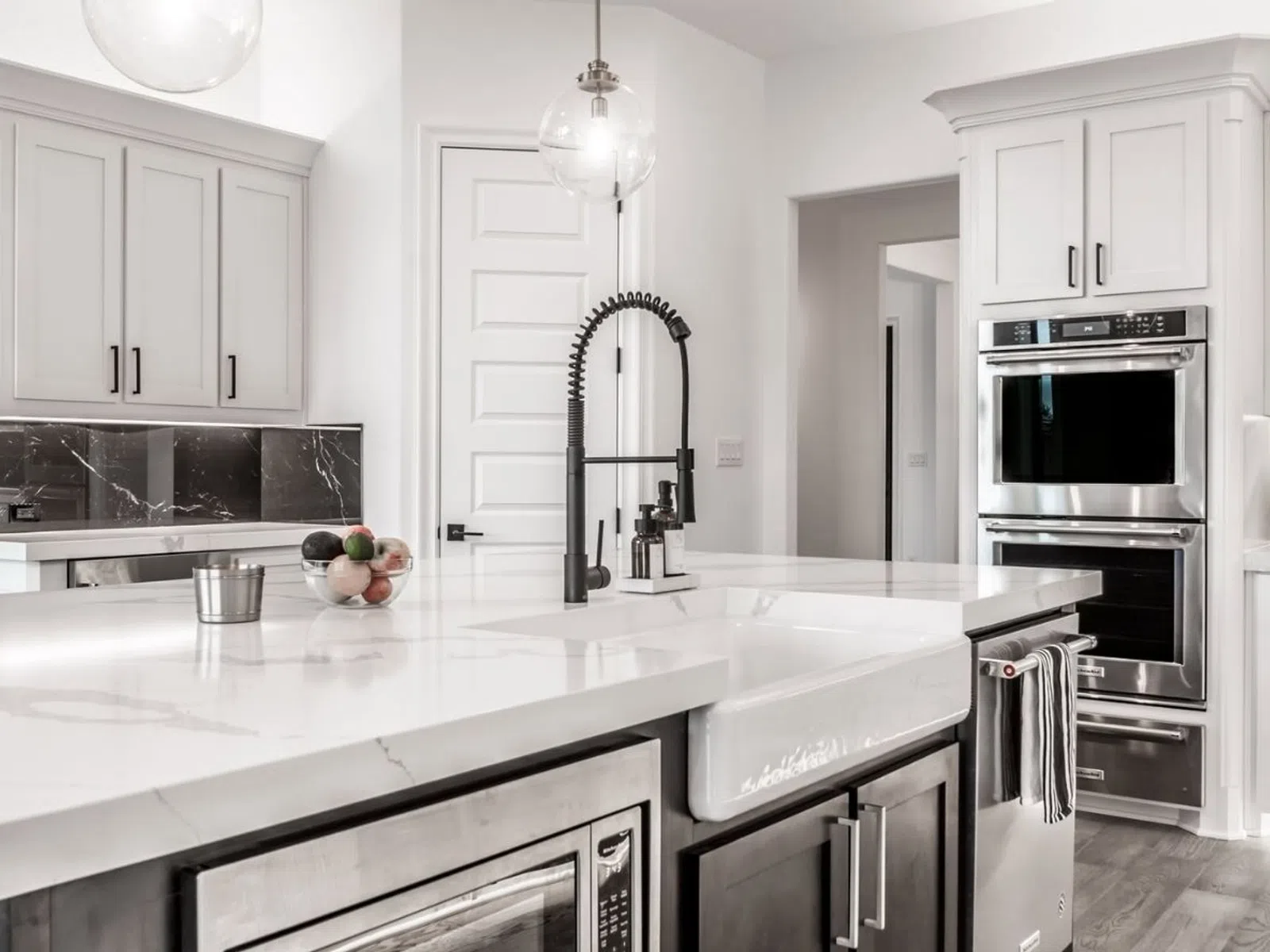 Modern white kitchen with marble countertops, farmhouse sink, stainless steel appliances, and pendant lighting.