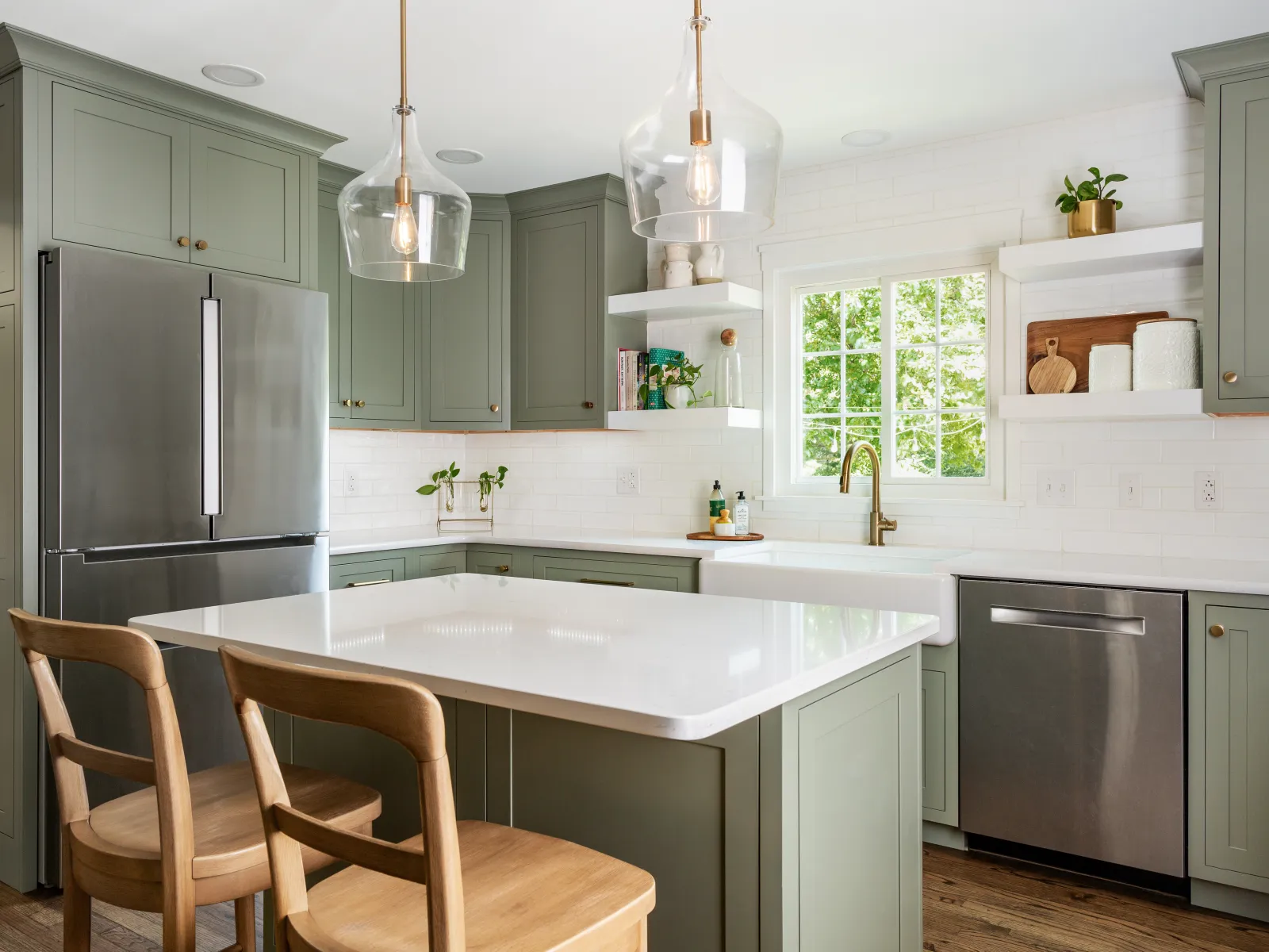 Modern kitchen with olive green cabinets, white countertops, wooden chairs, and stainless steel appliances under pendant lights