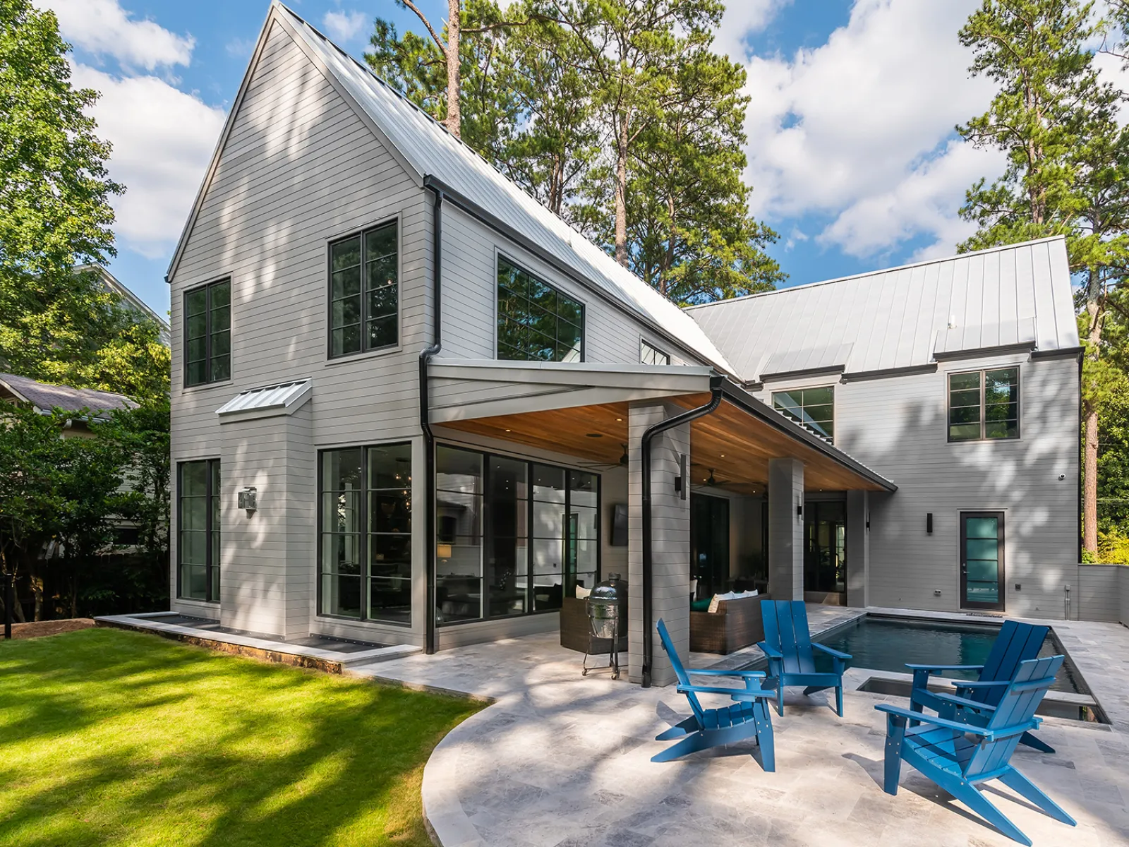 Modern two-story gray house with large windows, covered patio, blue outdoor chairs, and a swimming pool under sunny sky.