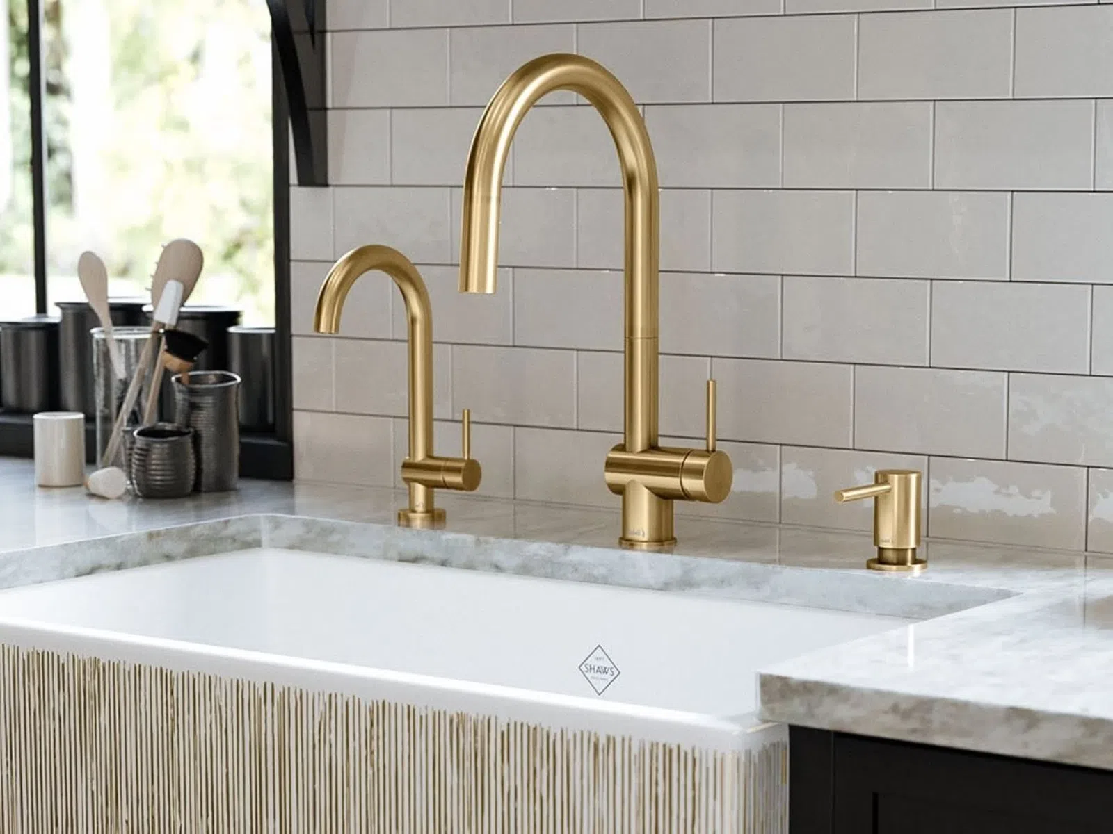 Modern kitchen sink with gold faucet, soap dispenser, and utensil holders on marble countertop under window.