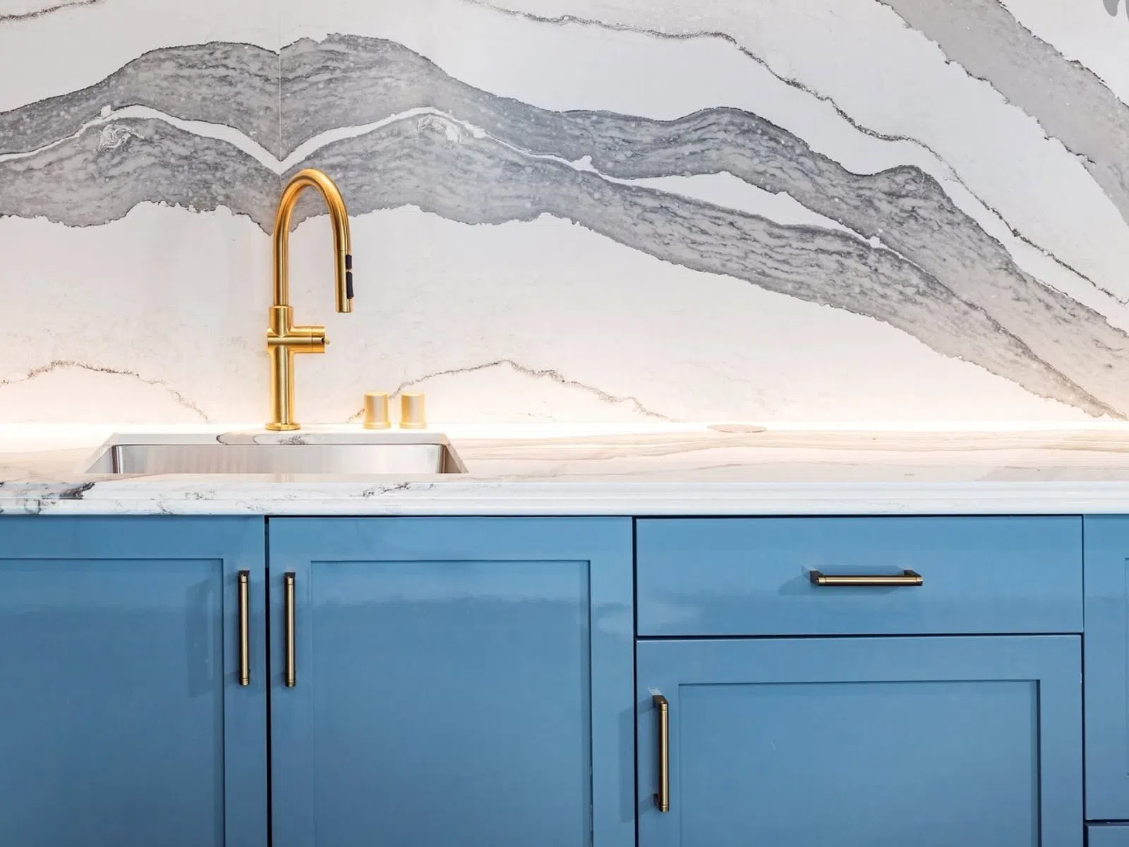 Modern kitchen sink with brass faucet set against blue cabinets and marble backsplash with gray veining