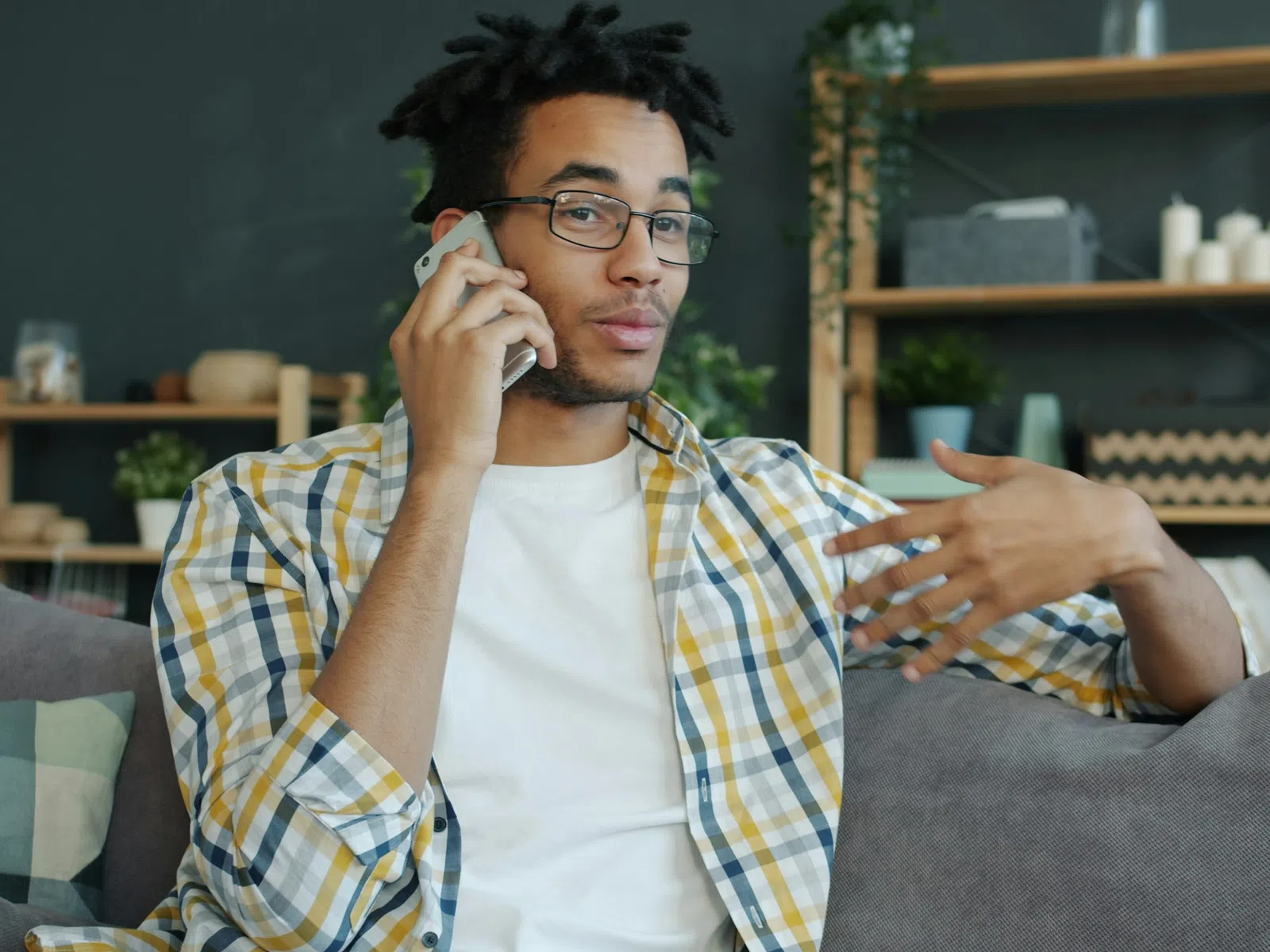 Young man with glasses wearing a plaid shirt talking on phone while sitting on couch in living room