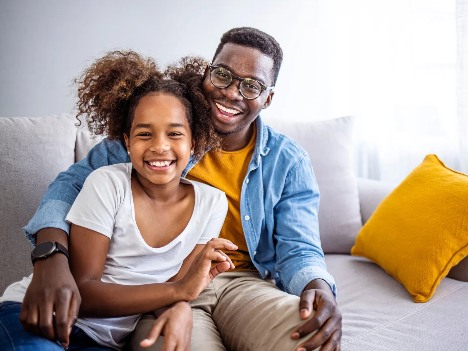 Smiling father and daughter sitting on a couch together in a bright living room with a yellow pillow nearby