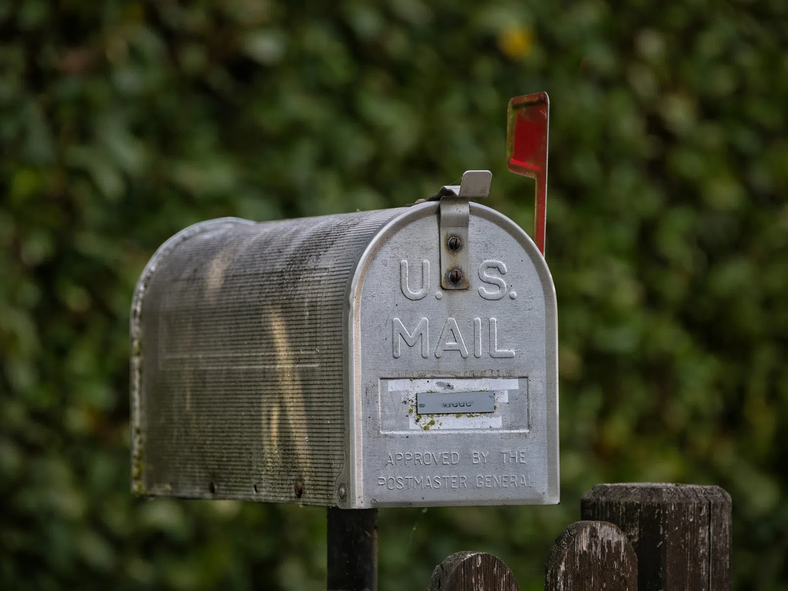 Rustic U.S. Mailbox with raised red flag on black post against blurred leafy background