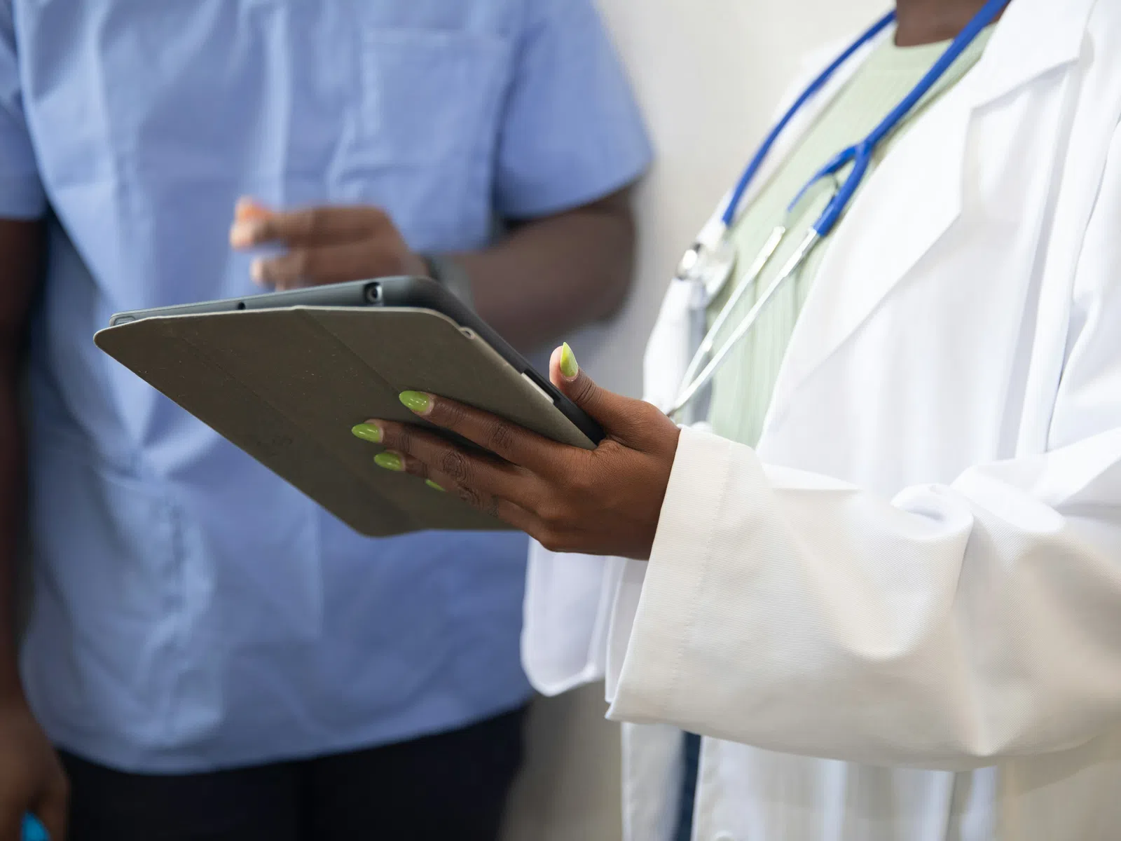 Doctor in white coat holding tablet with nurse in blue scrubs blurred in background