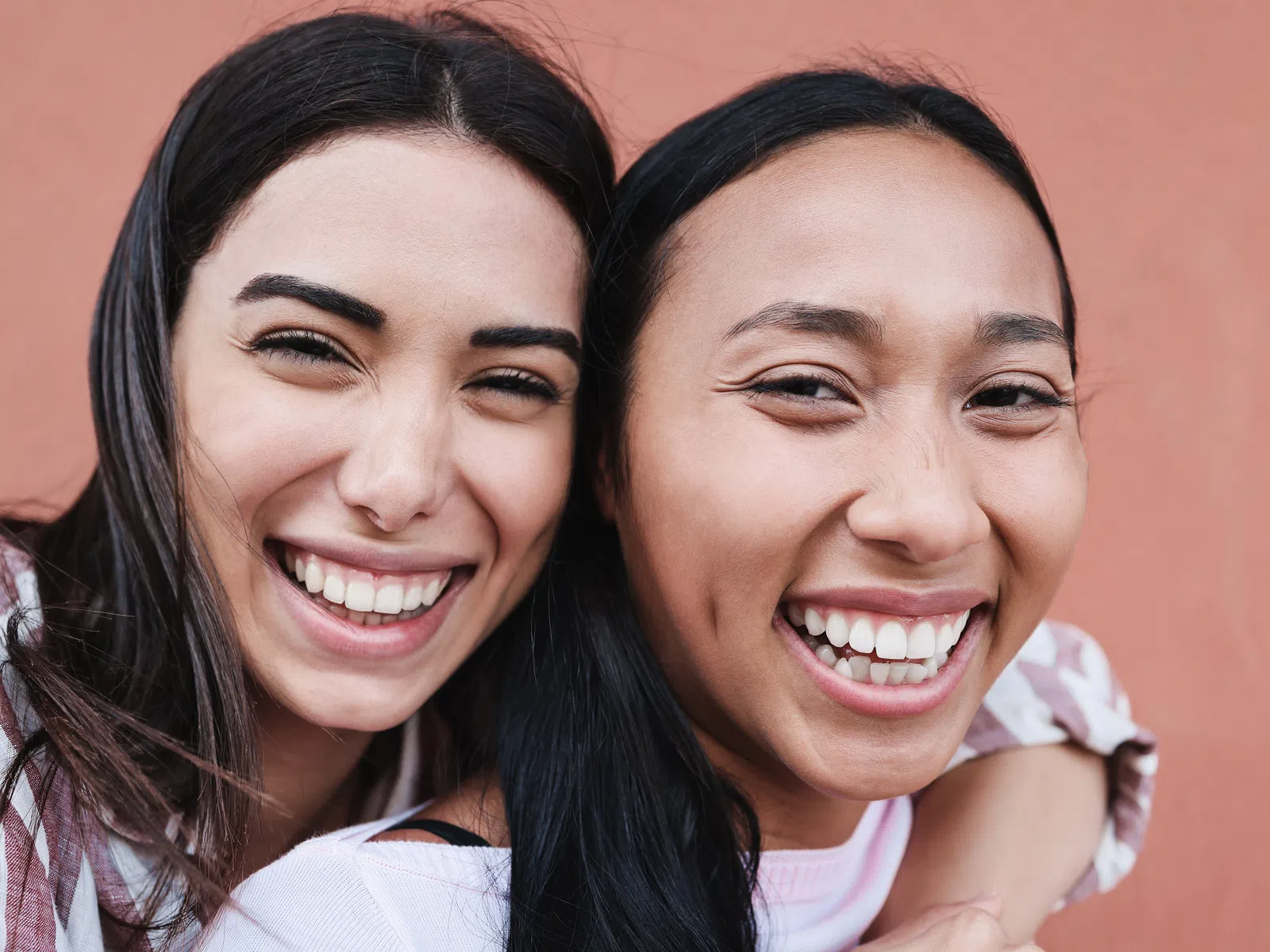 Two young women smiling and hugging against a warm peach background, showing friendship and happiness.