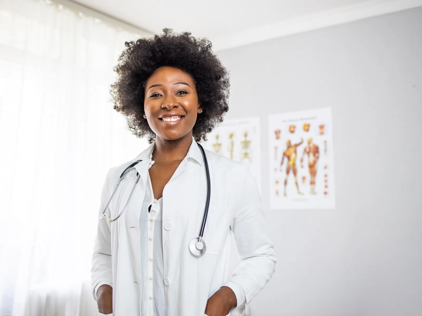 Smiling female doctor in white coat with stethoscope standing in bright medical office with anatomy charts.