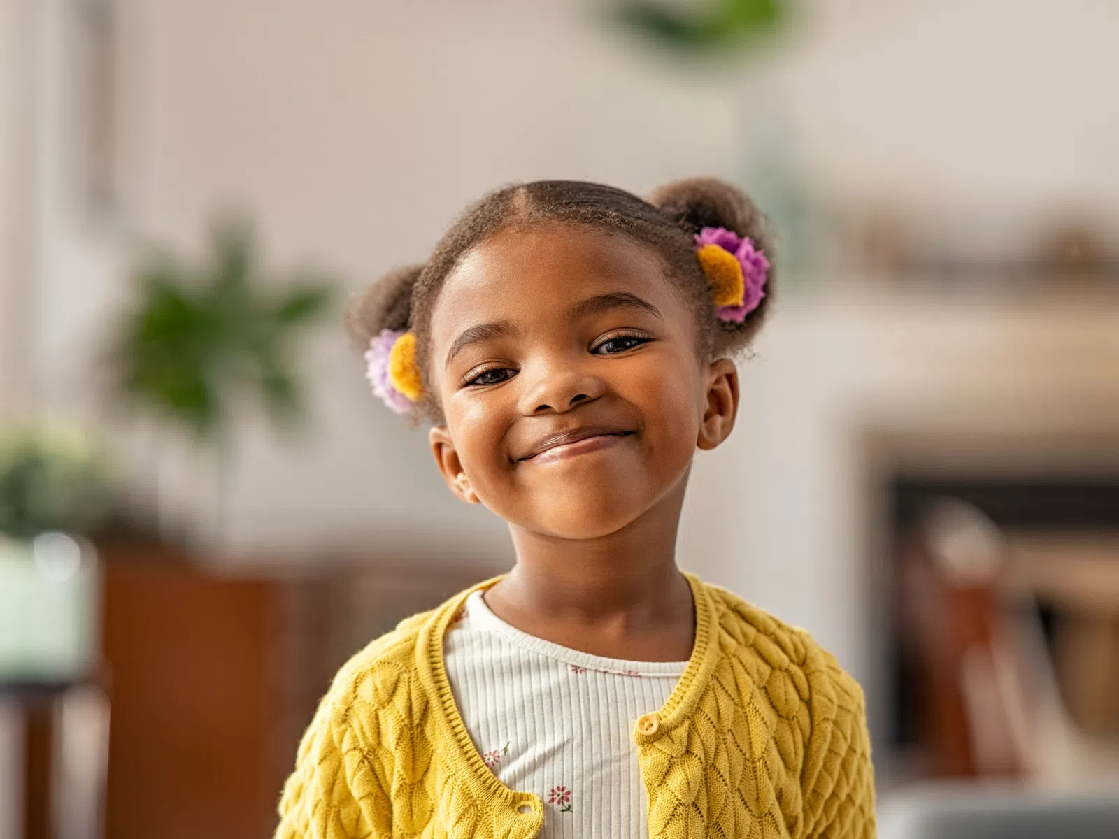 Smiling young girl with colorful hair ties wearing a yellow cardigan inside a cozy home setting