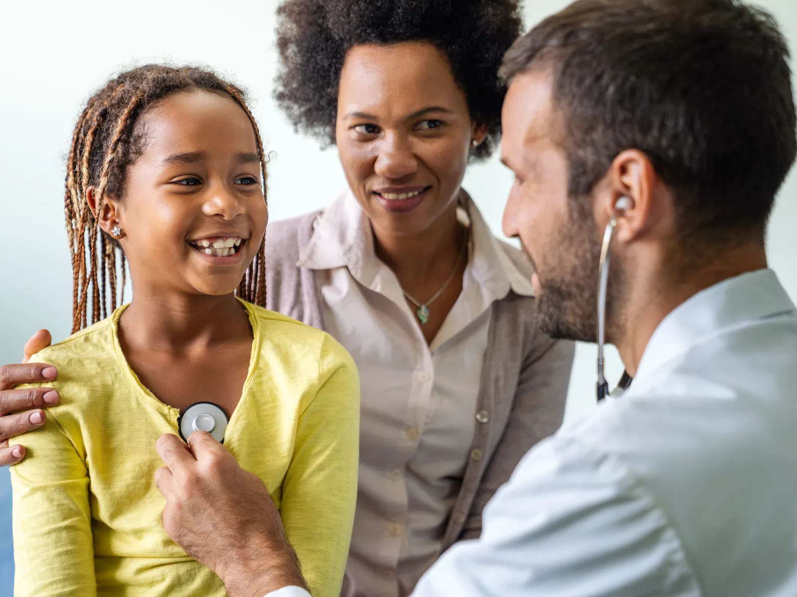 Doctor uses a stethoscope to check a smiling young girl's heartbeat while her mother looks on supportively.