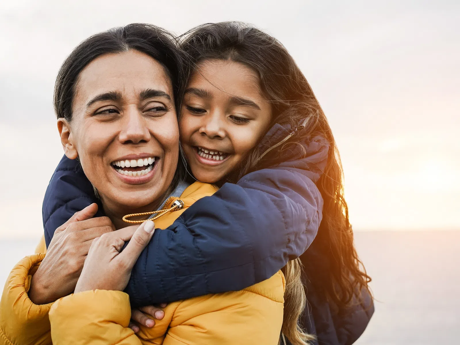 Mother and daughter smiling and hugging outdoors during sunset wearing warm jackets by the sea.