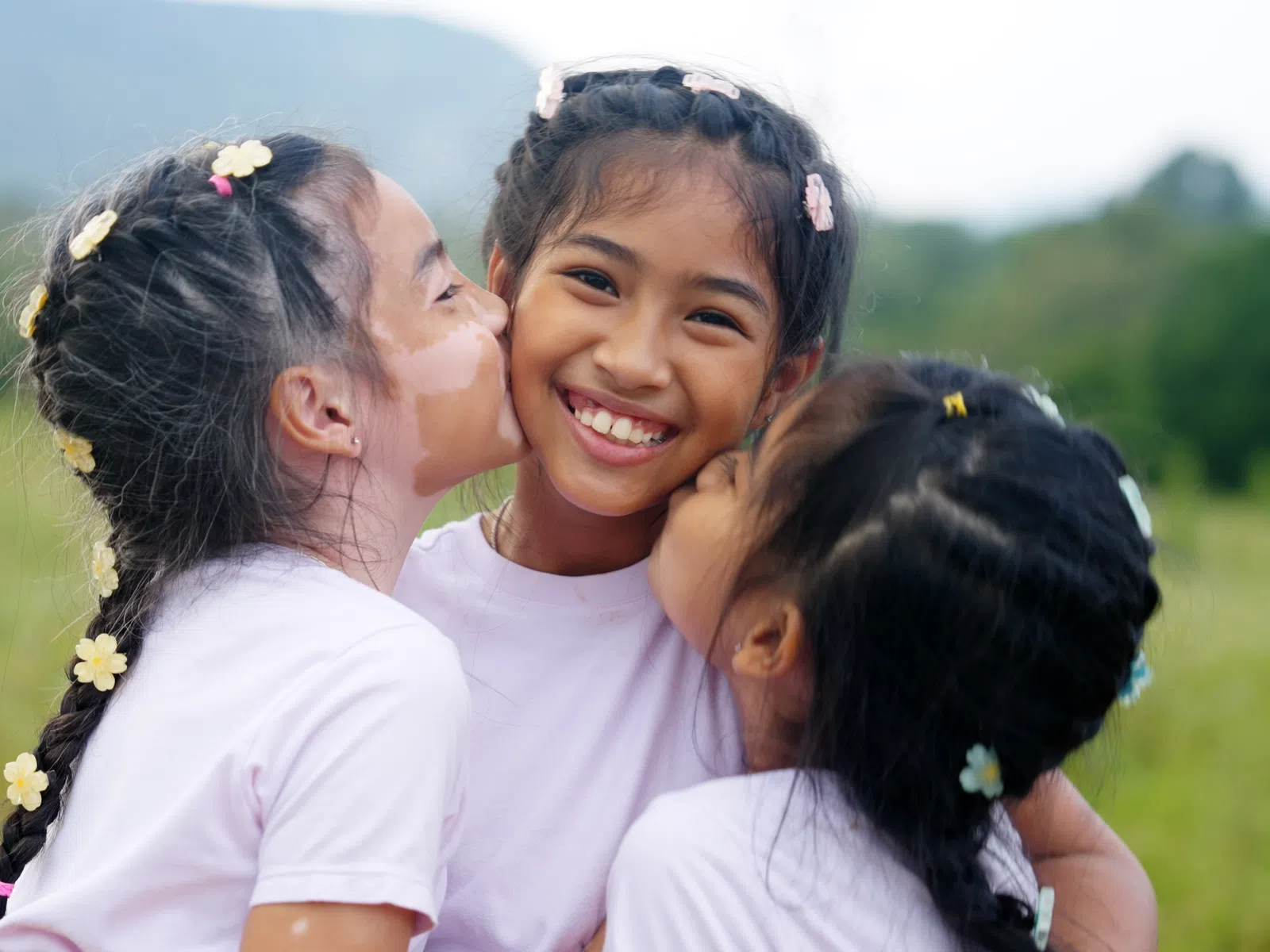 Three young girls smiling and hugging outdoors with flowers in their braided hair and a natural background.